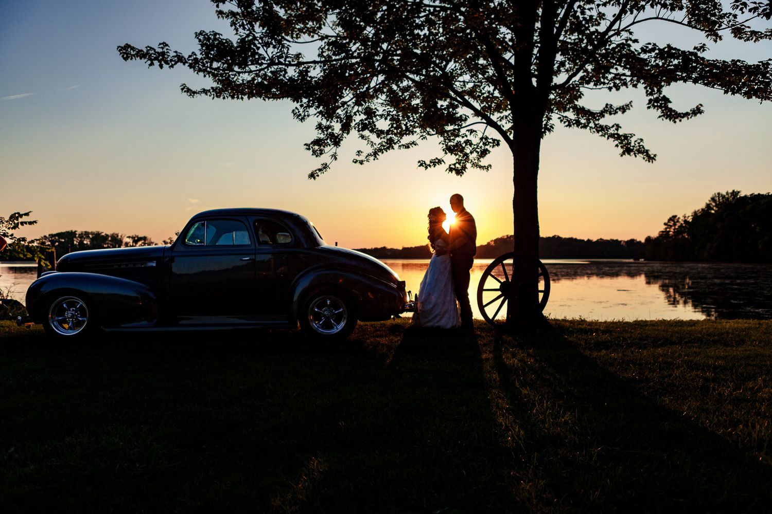 Romantic bride and groom silhouette at sunset beside a vintage car near a lake.