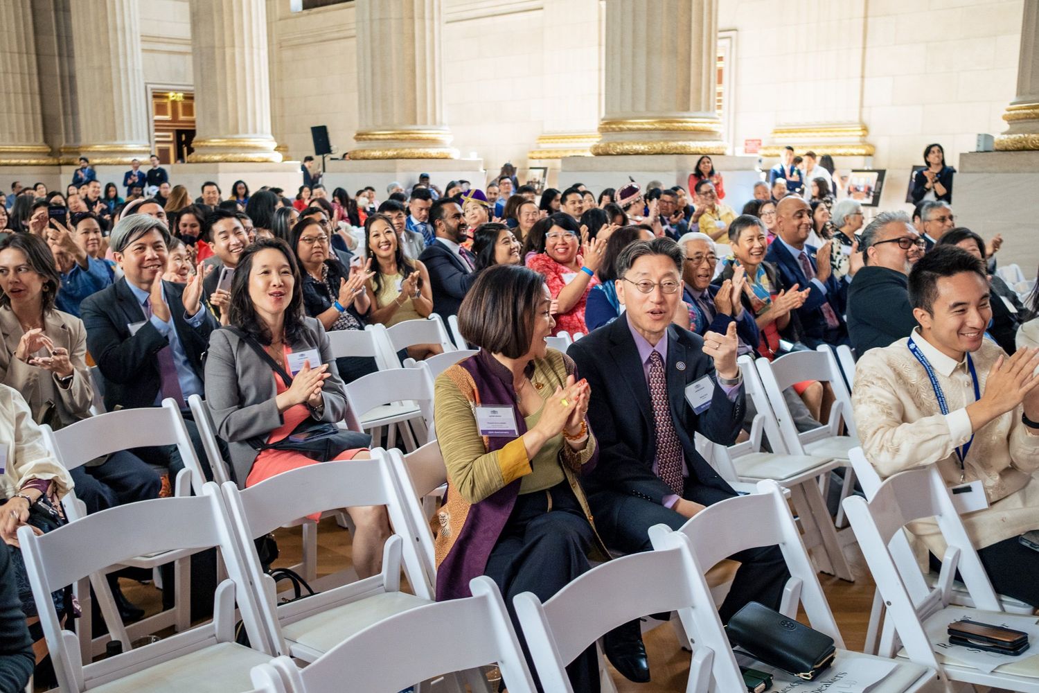 Diverse audience applauds at an elegant indoor ceremony with white chairs.
