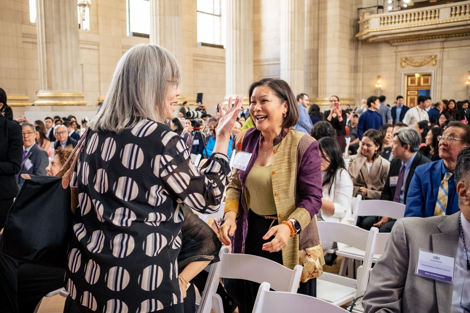 Two women greet each other warmly at a crowded professional conference in a grand hall.