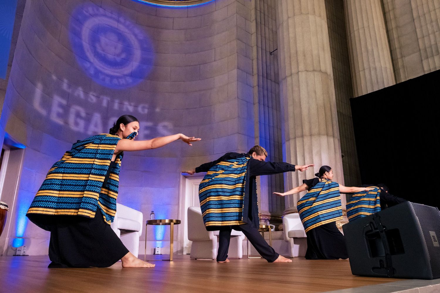 Performers in colorful striped robes dance on stage at a Lasting Legacies event inside a grand columned venue.