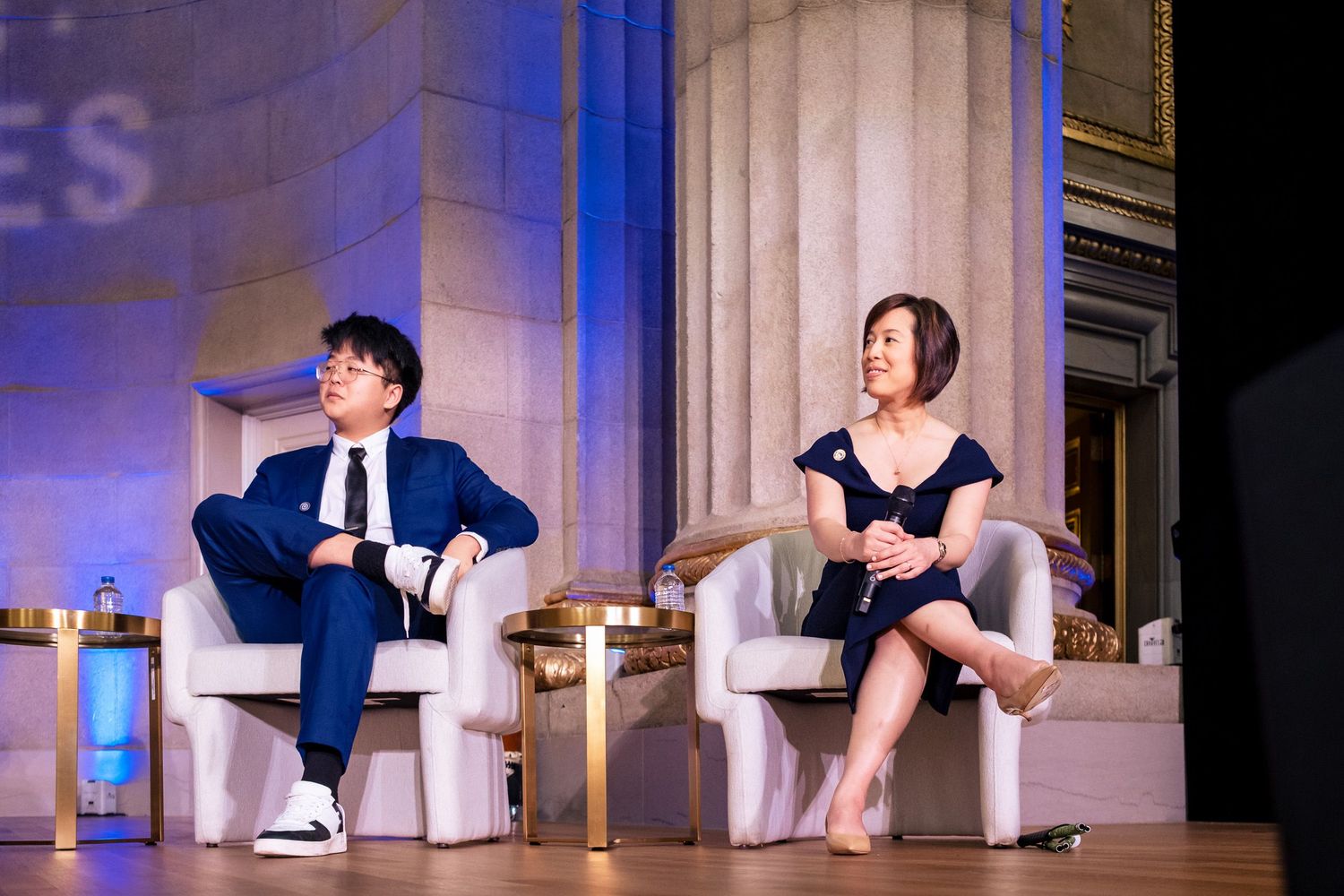 Two panelists seated in white chairs on a grand stage engaged in conversation at a formal event.