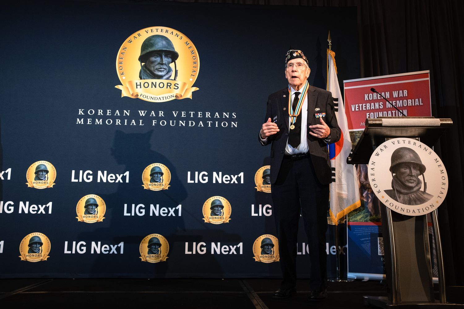 Veteran speaks at Korean War Veterans Memorial Foundation event with LIG Nex1 sponsored backdrop.