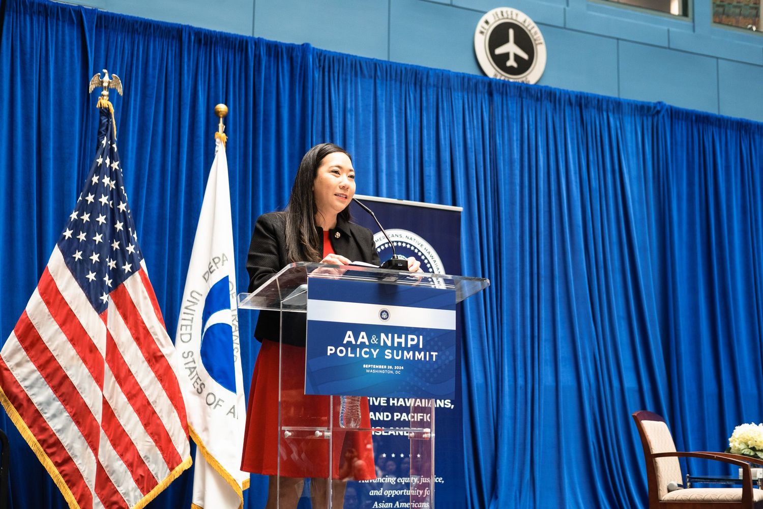 Asian American woman speaks at podium during AA&NHPI Policy Summit with US flag and organizational banner.