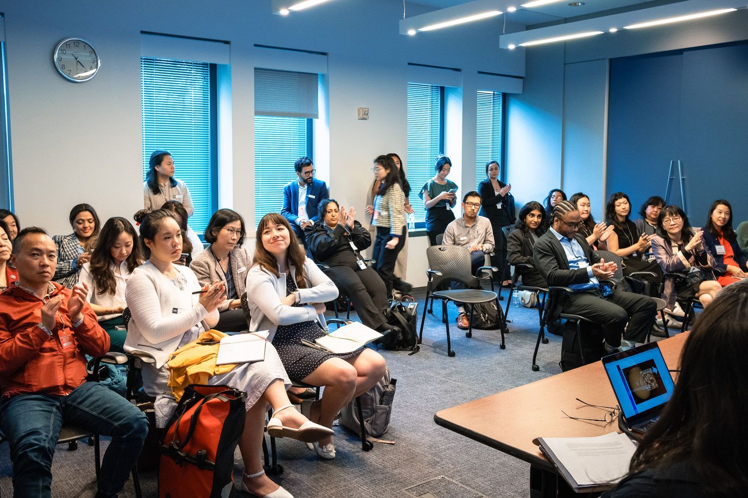 A diverse group of students attends a classroom lecture in a modern blue-accented university seminar room.