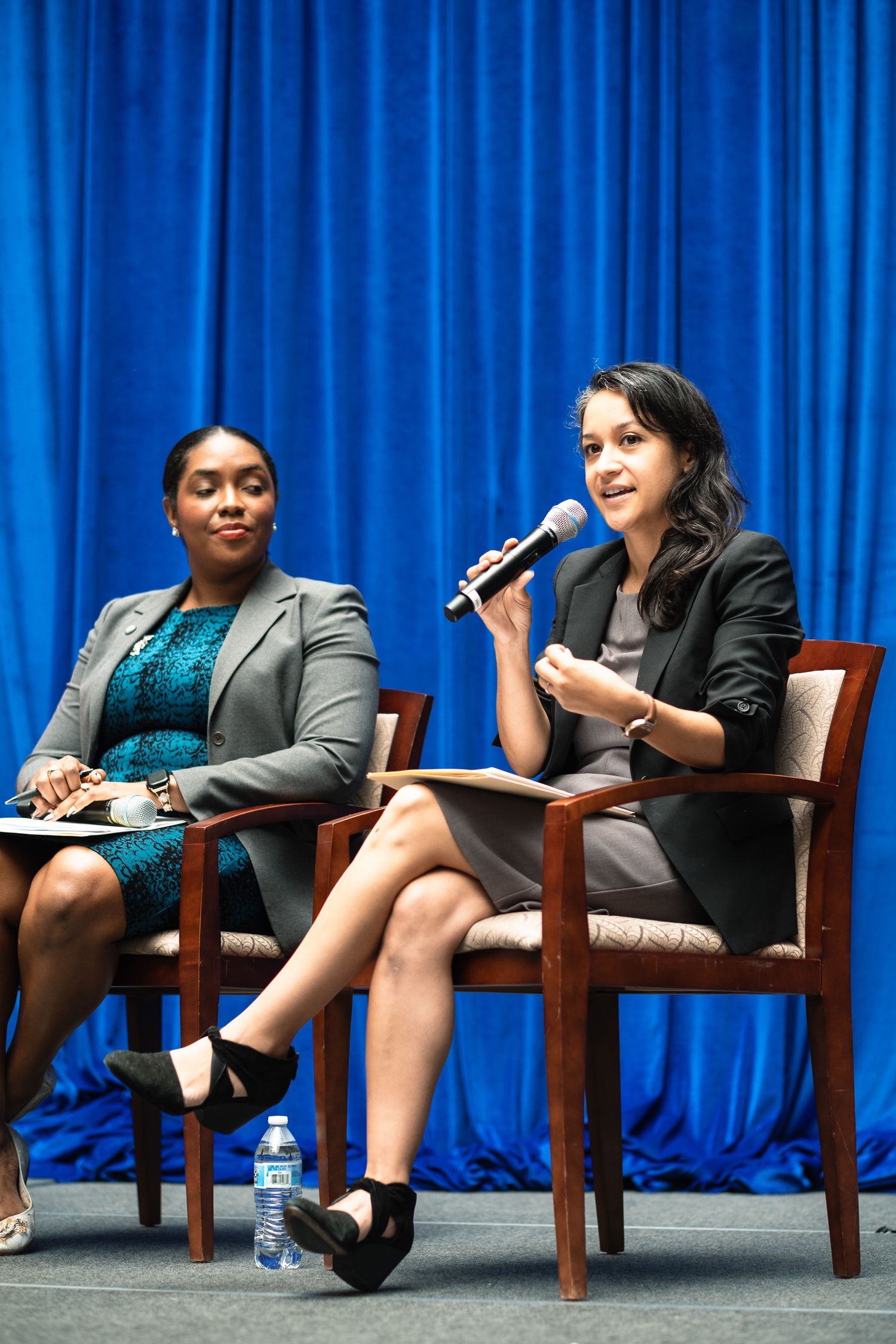 Two professional women in blazers sit in chairs during a panel discussion, one speaking into a microphone before a blue curtain.