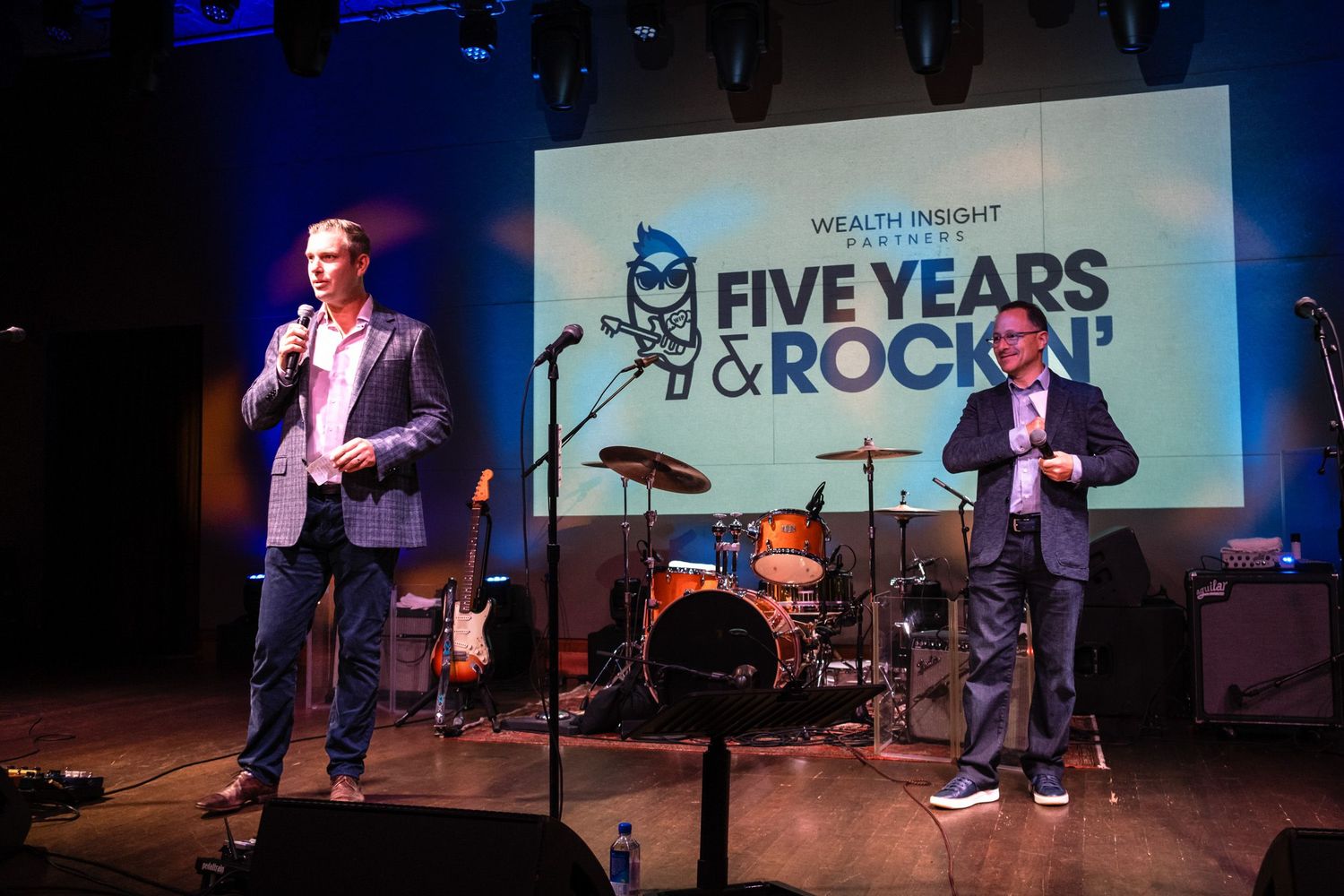Two men on stage at Wealth Insight Partners Five Years & Rockin' anniversary event with band equipment behind them.