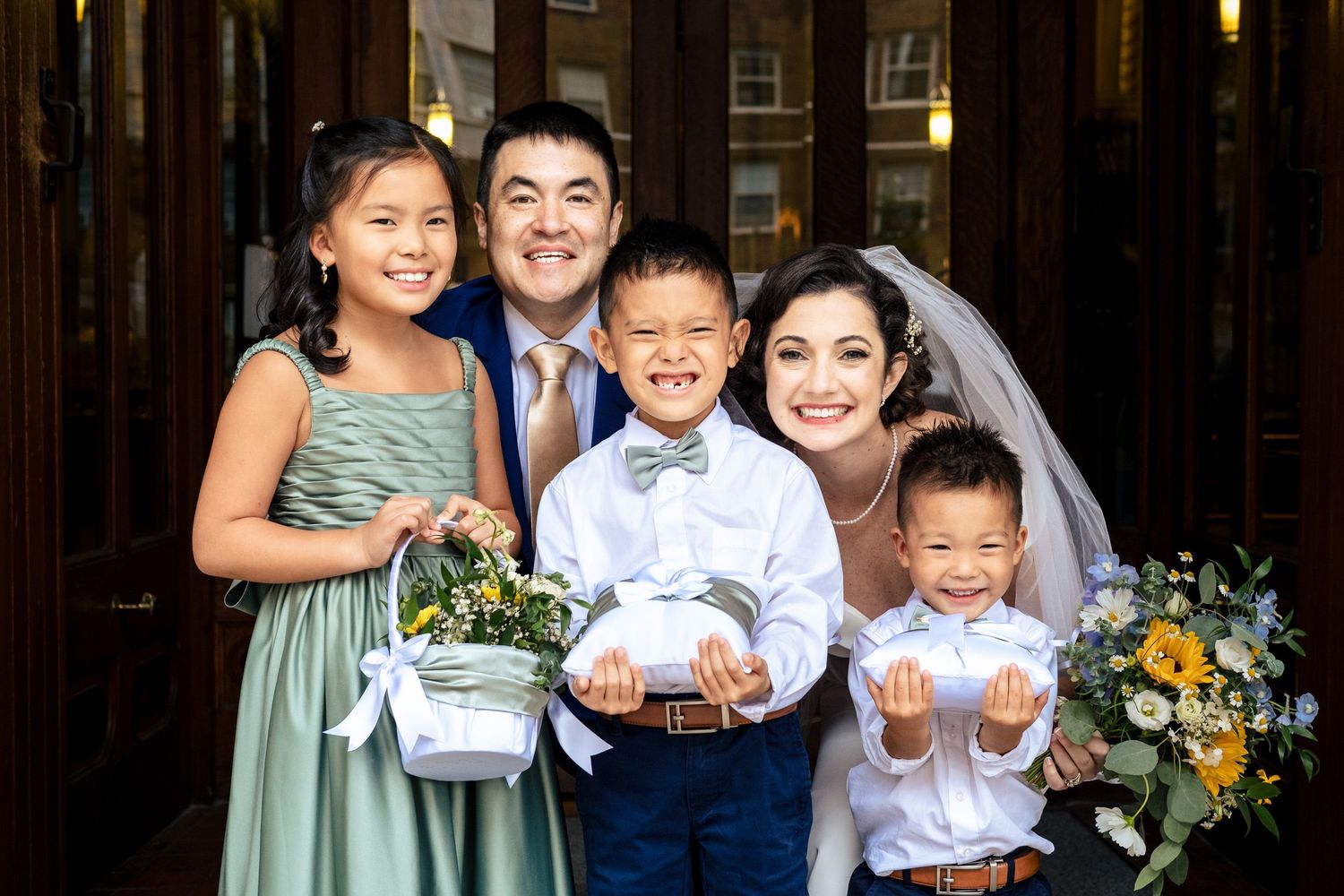 Bride and groom posing with three children in sage green and navy attire holding flowers at church entrance.