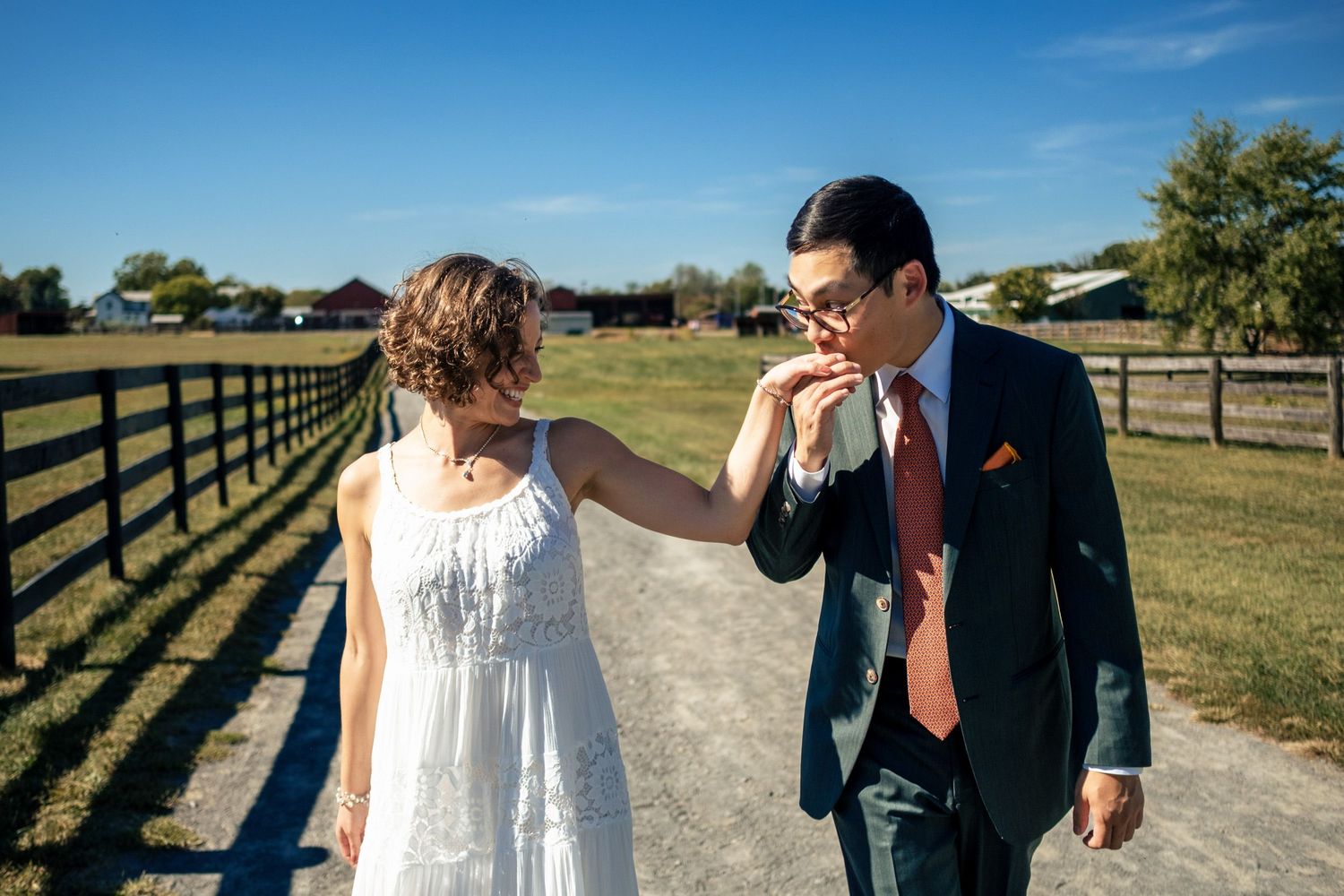 A groom gallantly kisses his bride's hand while strolling along a farm path.