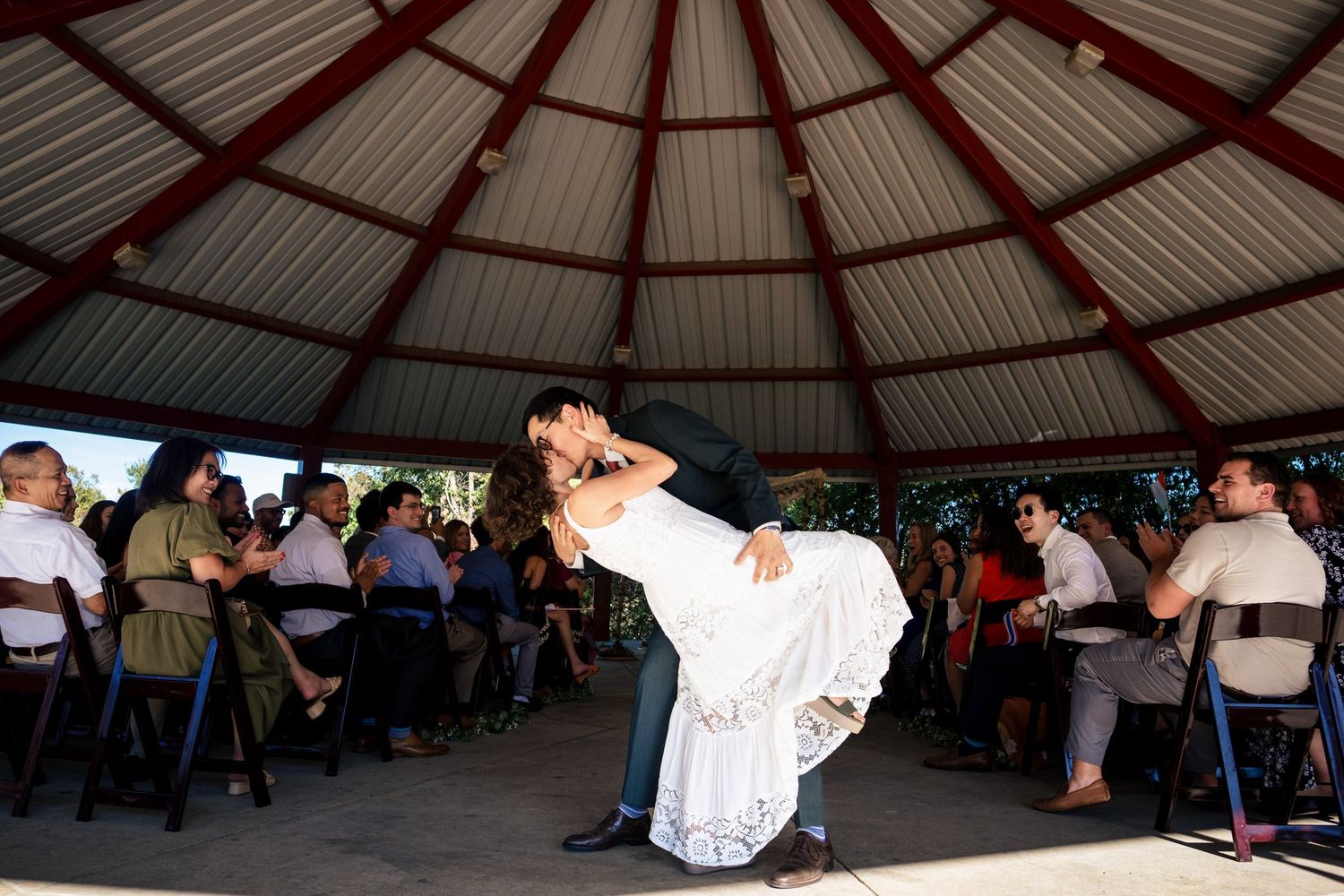 Groom dips bride during first dance in outdoor pavilion surrounded by wedding guests.