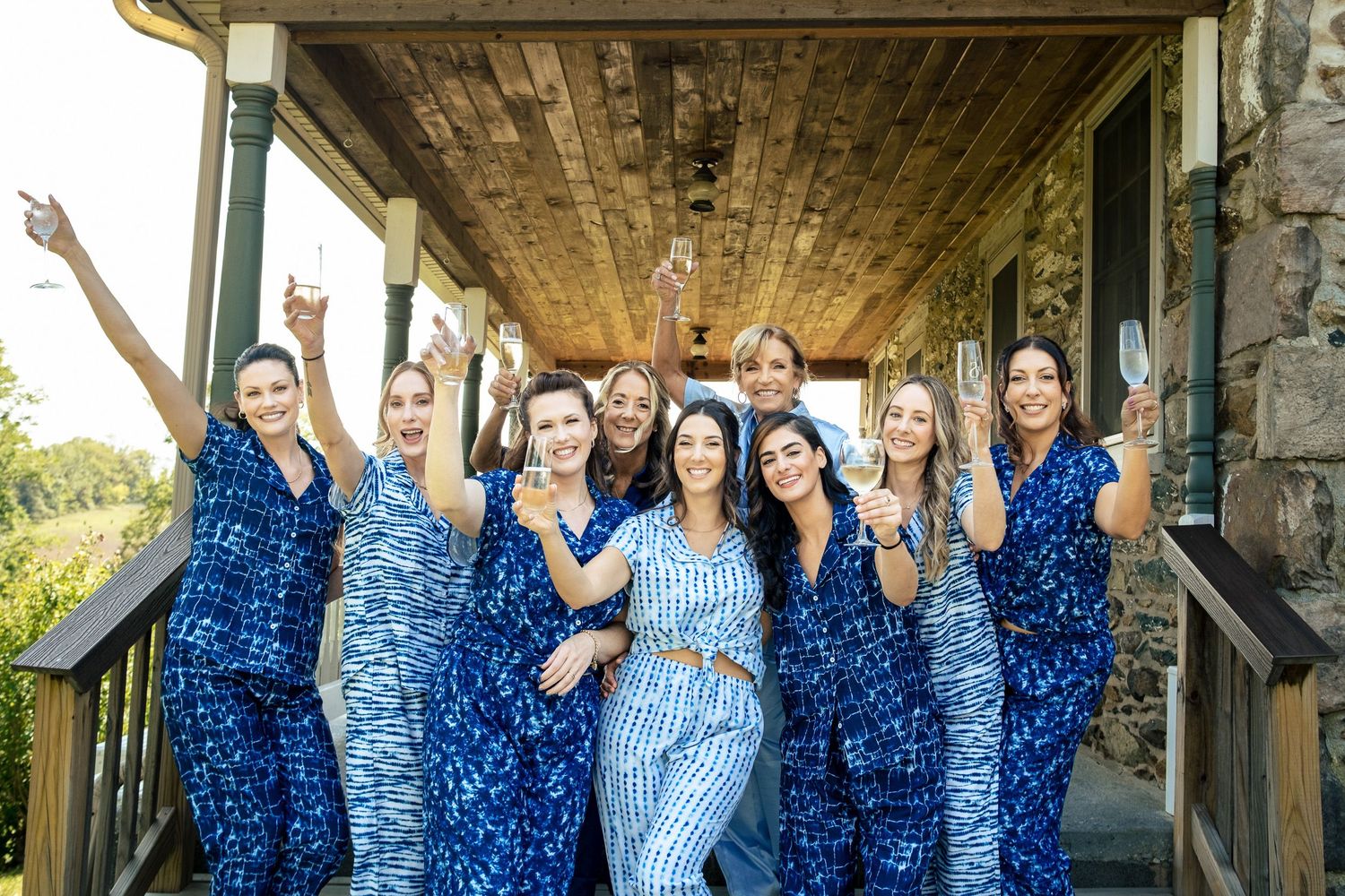 Bride and bridesmaids in matching blue pajamas celebrate together on a rustic stone porch.
