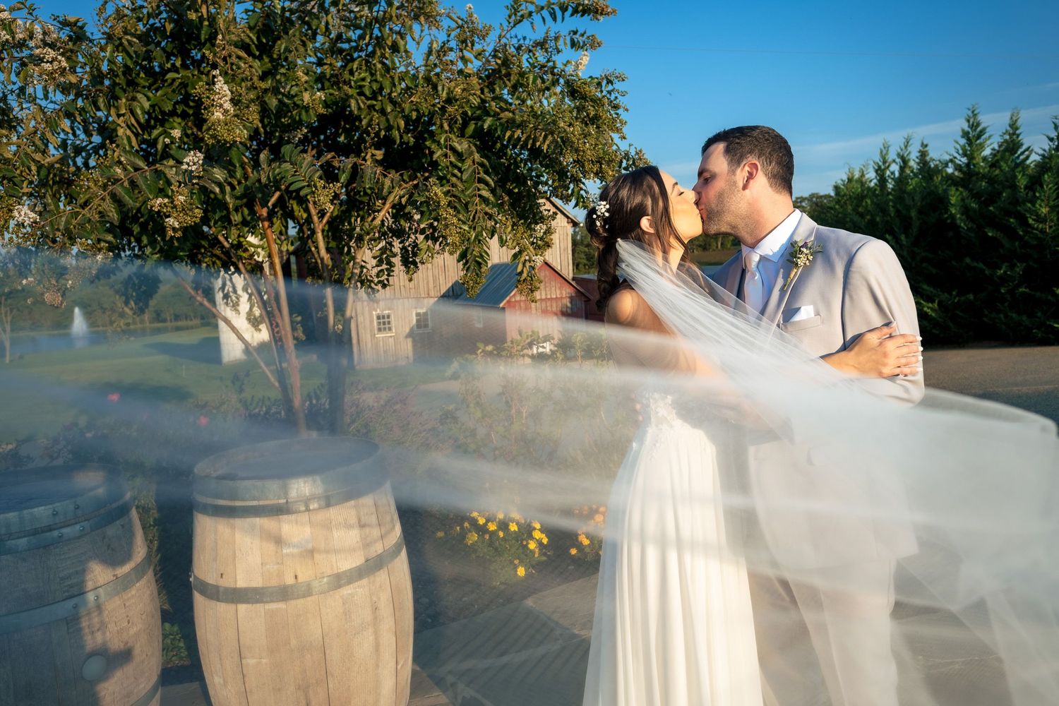 Bride and groom share a kiss as her flowing veil sweeps across the frame beside rustic wooden barrels.