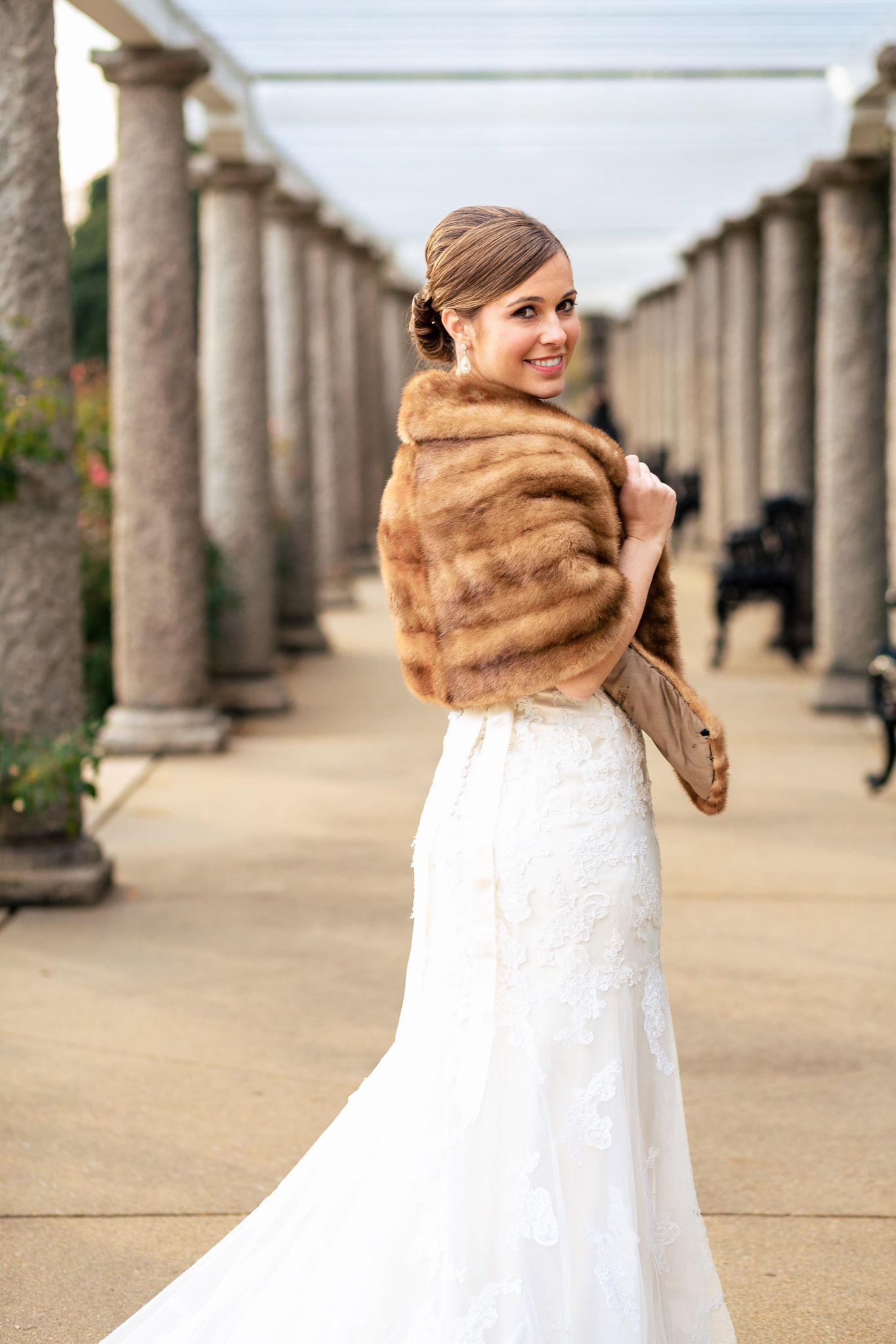 Bride in white lace gown wearing vintage fur stole between stone columns smiling.