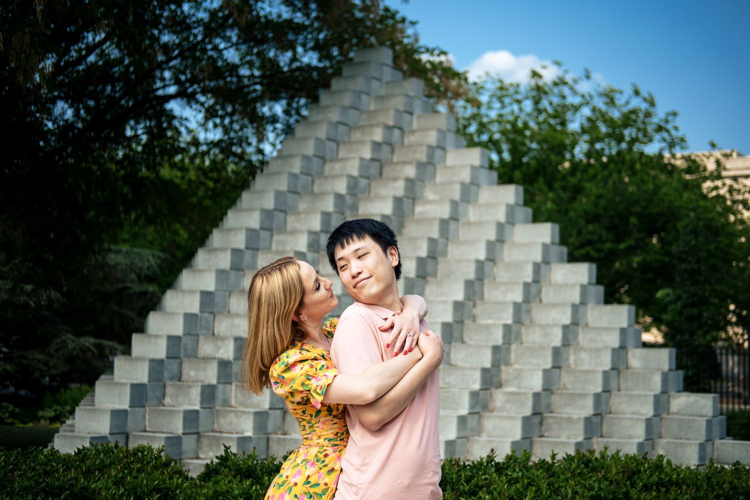 Happy couple embracing and smiling in front of a geometric pyramid sculpture in a sunny park.