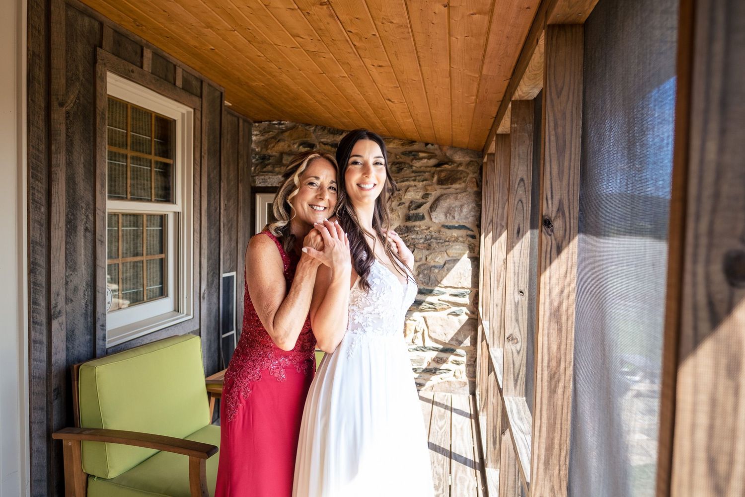Bride in white wedding dress posing with smiling bridesmaid in red dress on rustic porch.