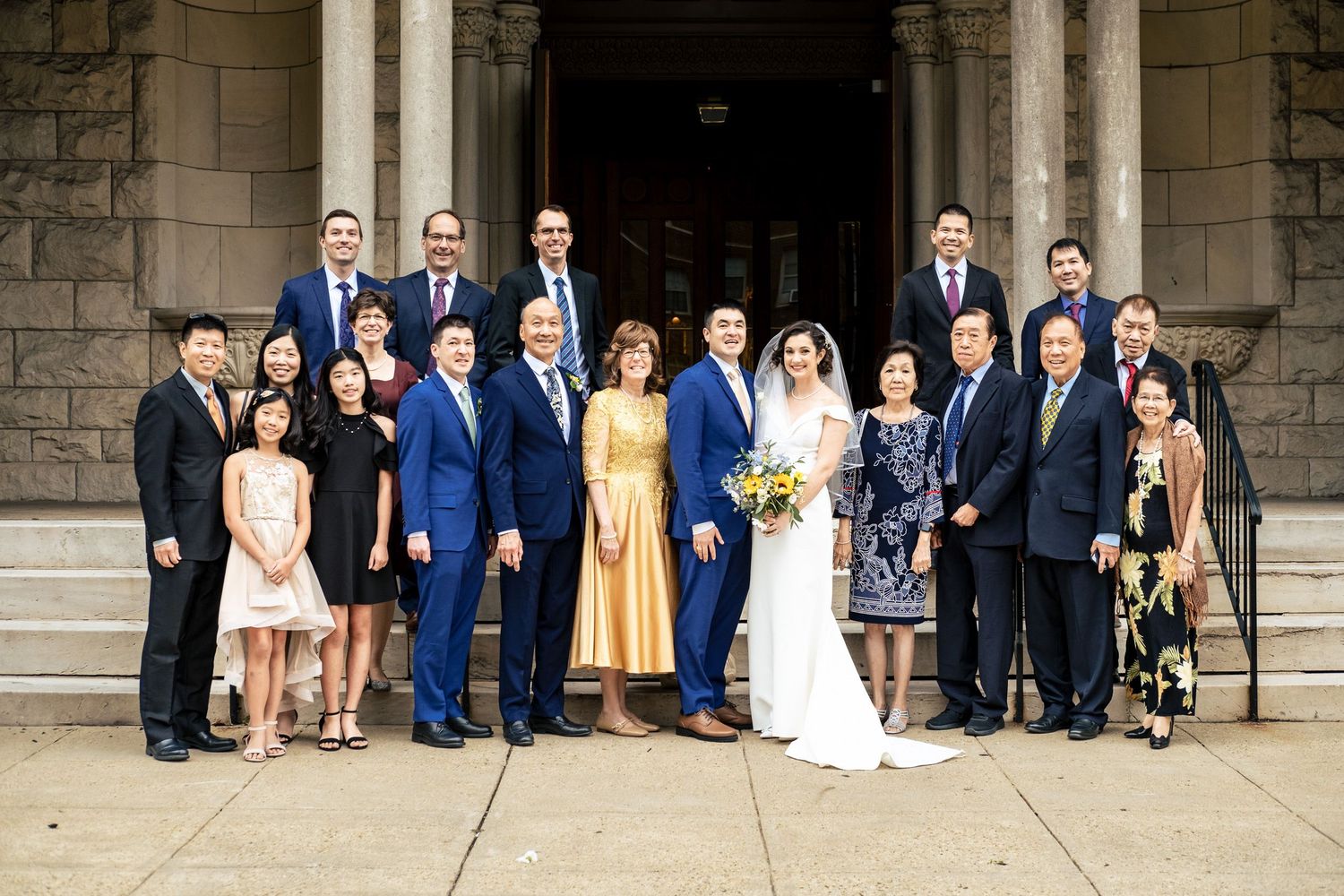 Large wedding party group portrait on stone church steps with bride in white gown holding yellow sunflower bouquet.