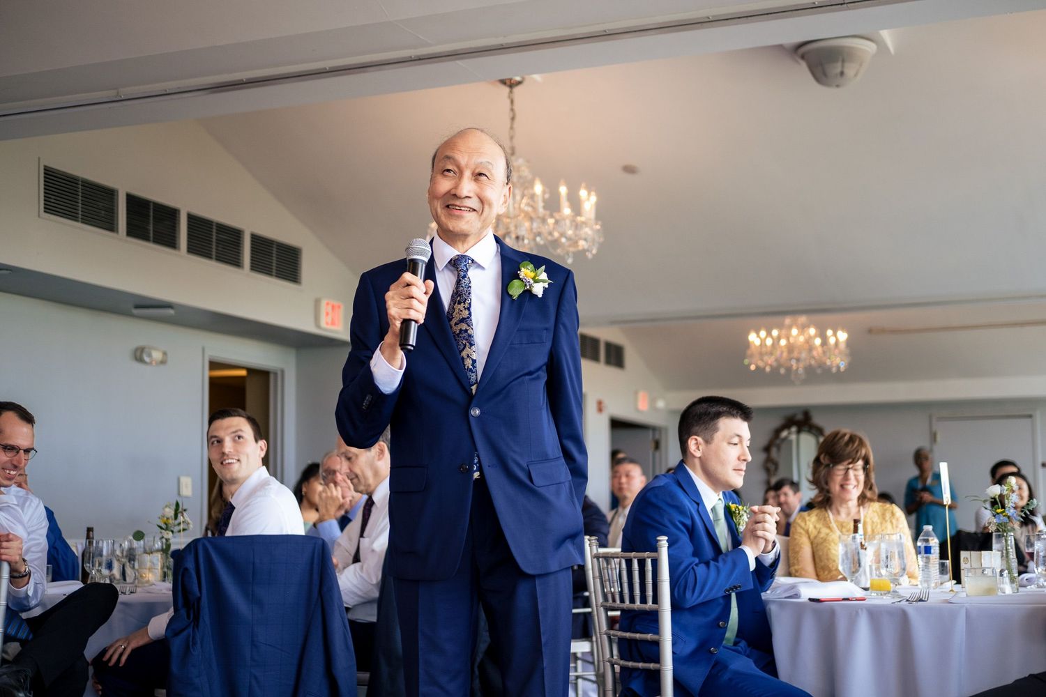 Man in blue suit delivers a wedding speech with a microphone in an elegant chandelier-lit reception hall.
