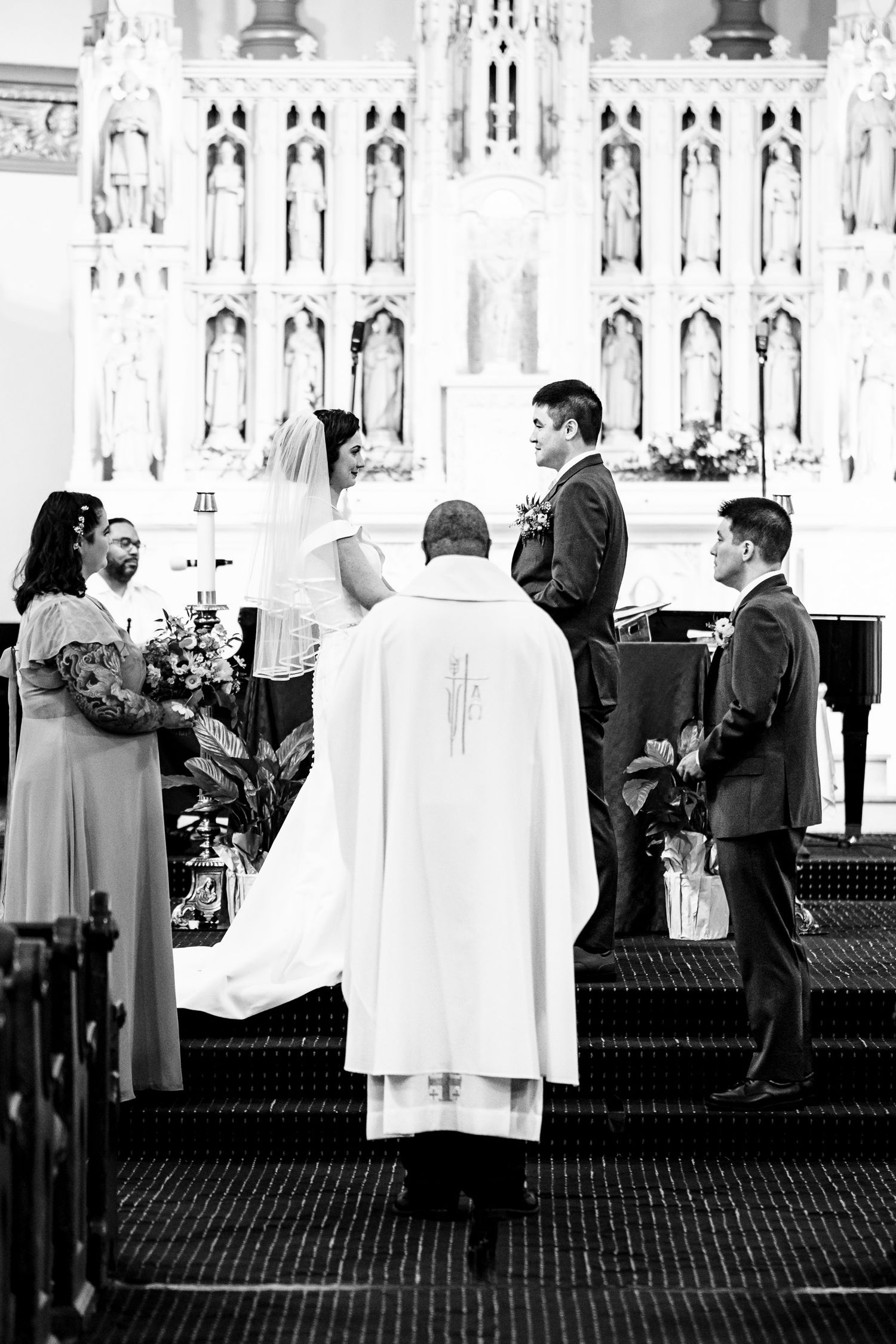 Black and white photo of bride and groom exchanging vows before a priest at a church altar.