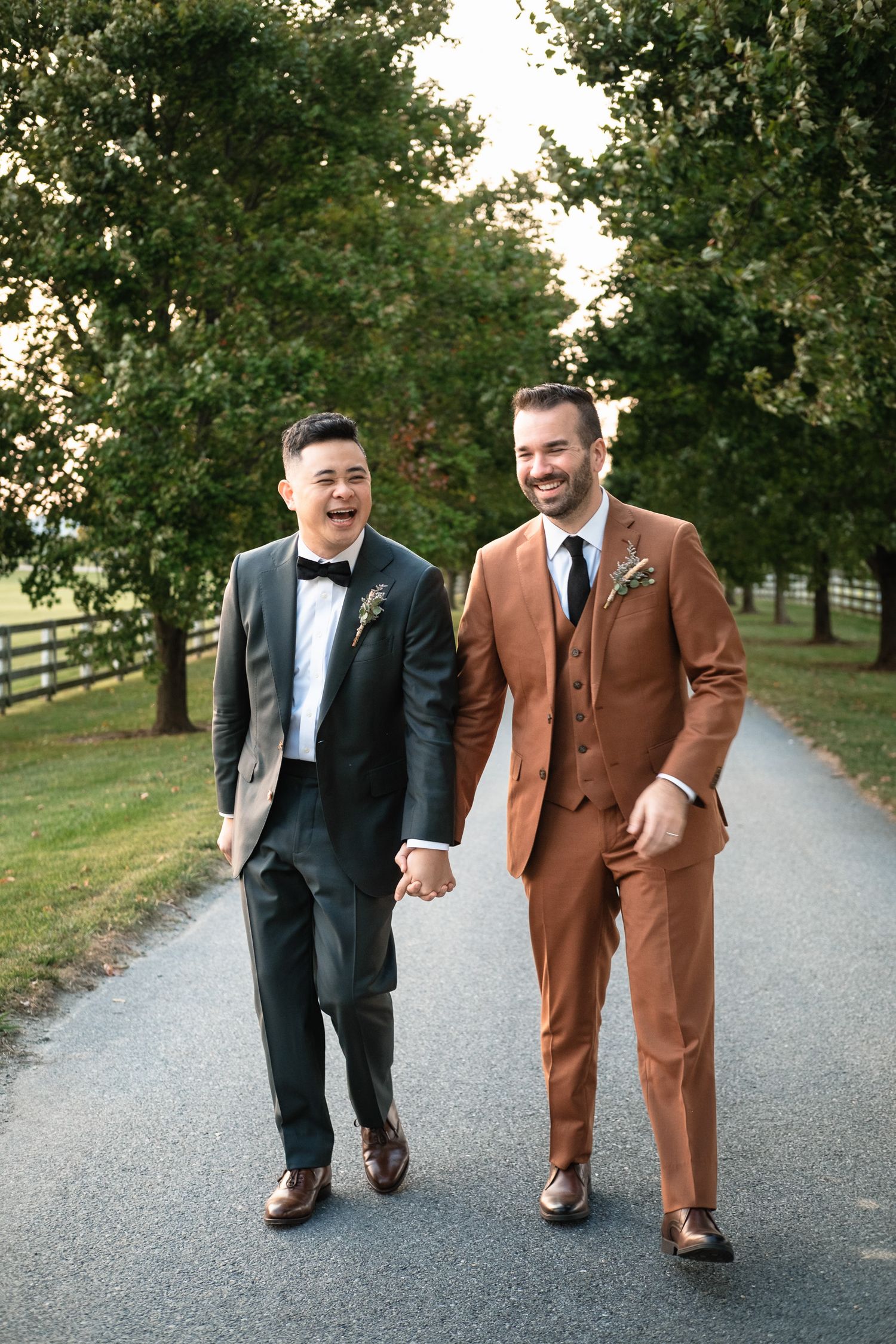 Two grooms hold hands and laugh while walking down a tree-lined path on their wedding day.