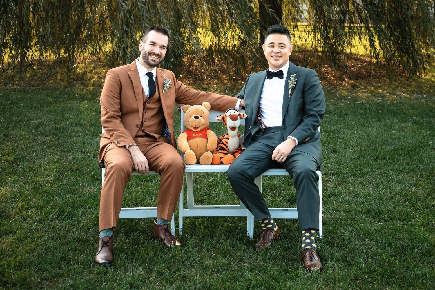 Two grooms in suits sit on a white bench with Winnie the Pooh teddy bear at outdoor wedding.