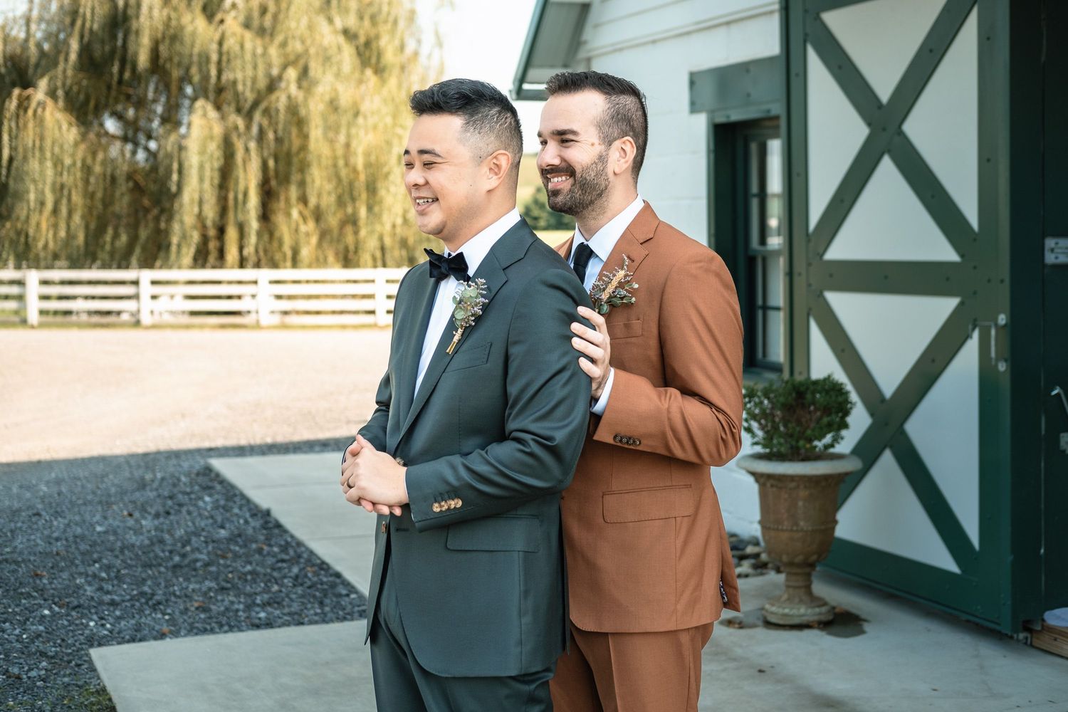 Two grooms share a tender first look moment outside a white barn wedding venue.