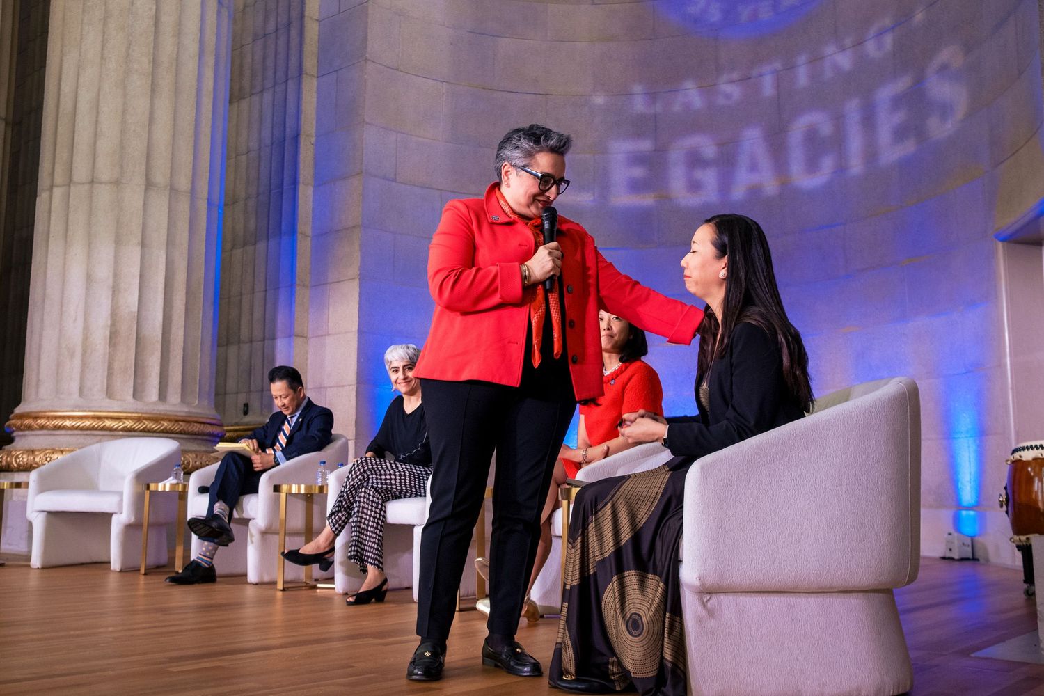 Speaker in red jacket holds microphone near seated woman at Lasting Legacies event in elegant venue.