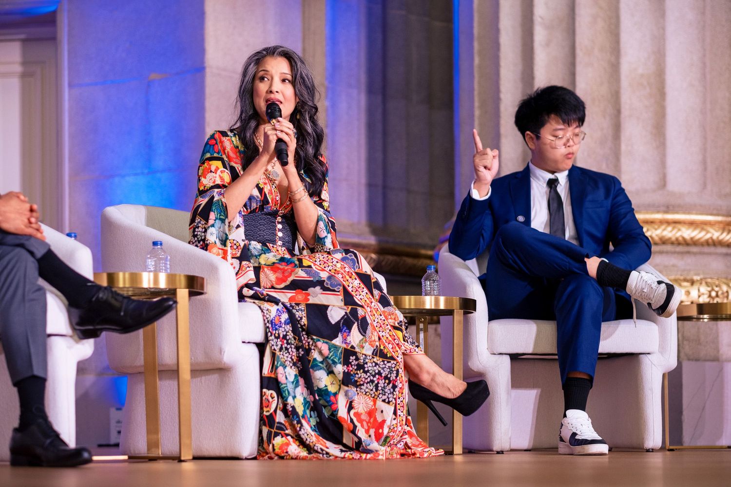 Woman in floral dress speaks at panel event beside man in blue suit in elegant venue.