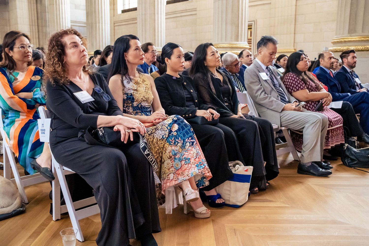 Diverse group of people seated in rows at a formal event inside an elegant hall with classical architecture.