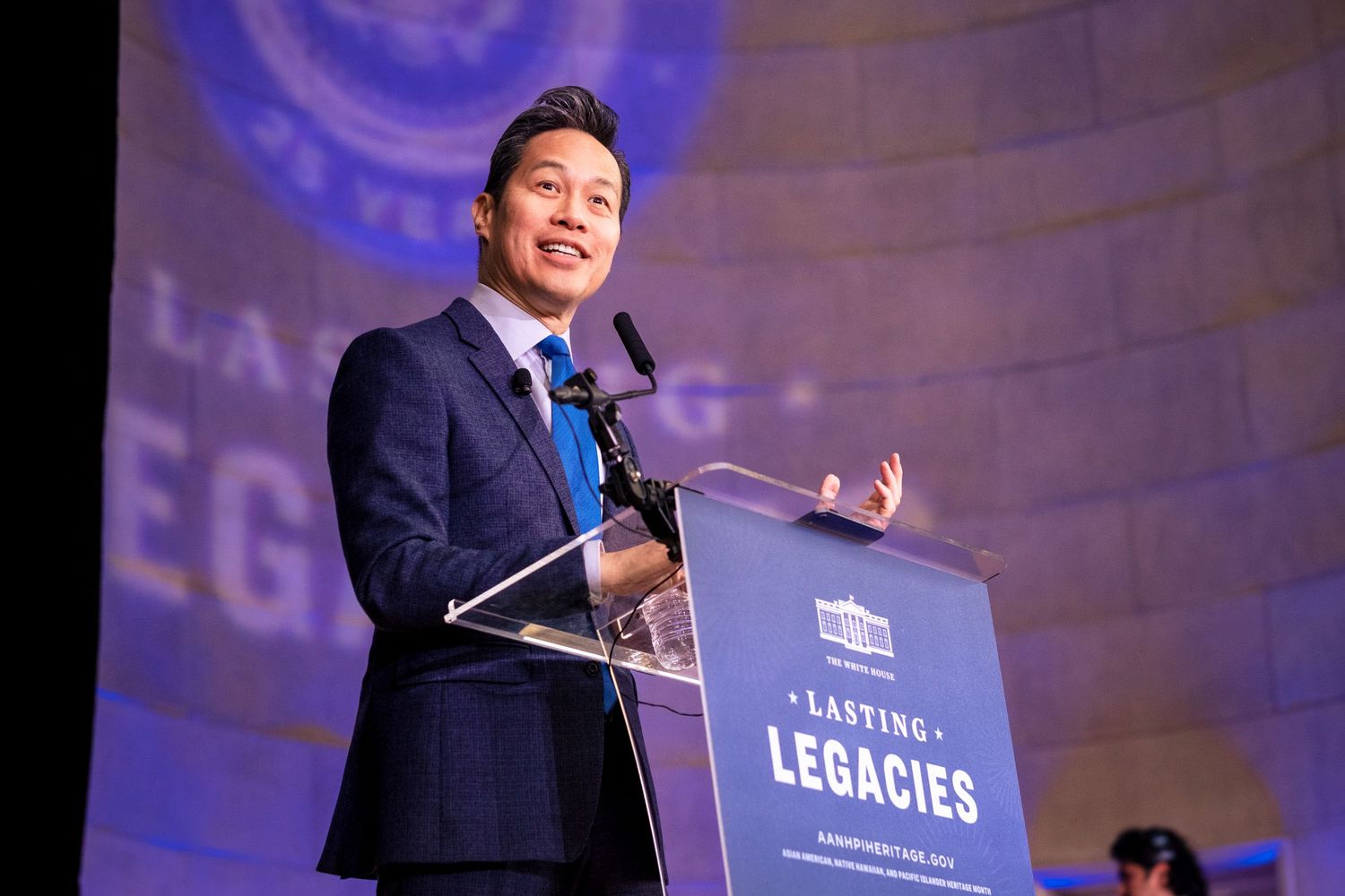 Man in blue suit speaking at podium during Lasting Legacies event on stage with purple backdrop.