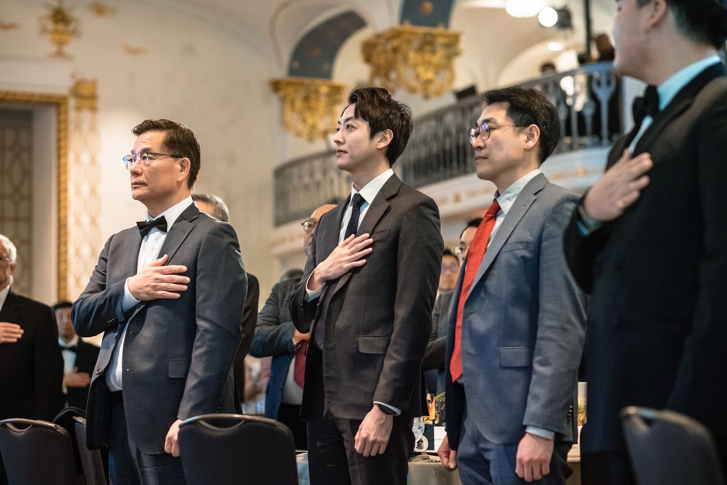 Men in formal attire stand with hands on hearts during a ceremony in an ornate hall.