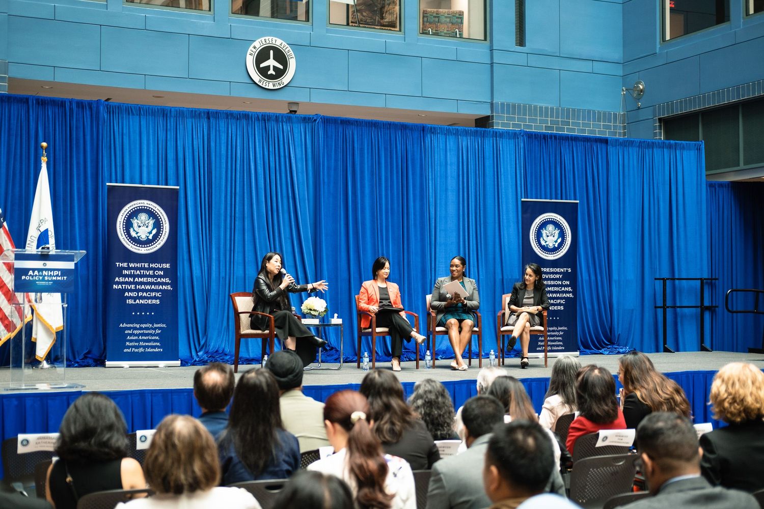 Panel discussion on stage with blue curtains at a formal government event with audience.