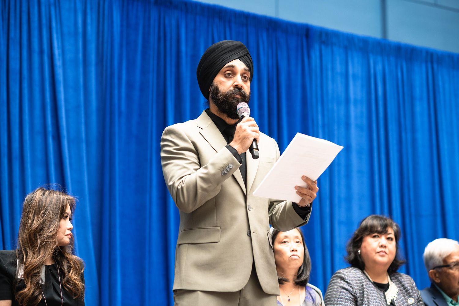Man in turban speaks at podium with microphone, holding papers at formal event with seated audience.