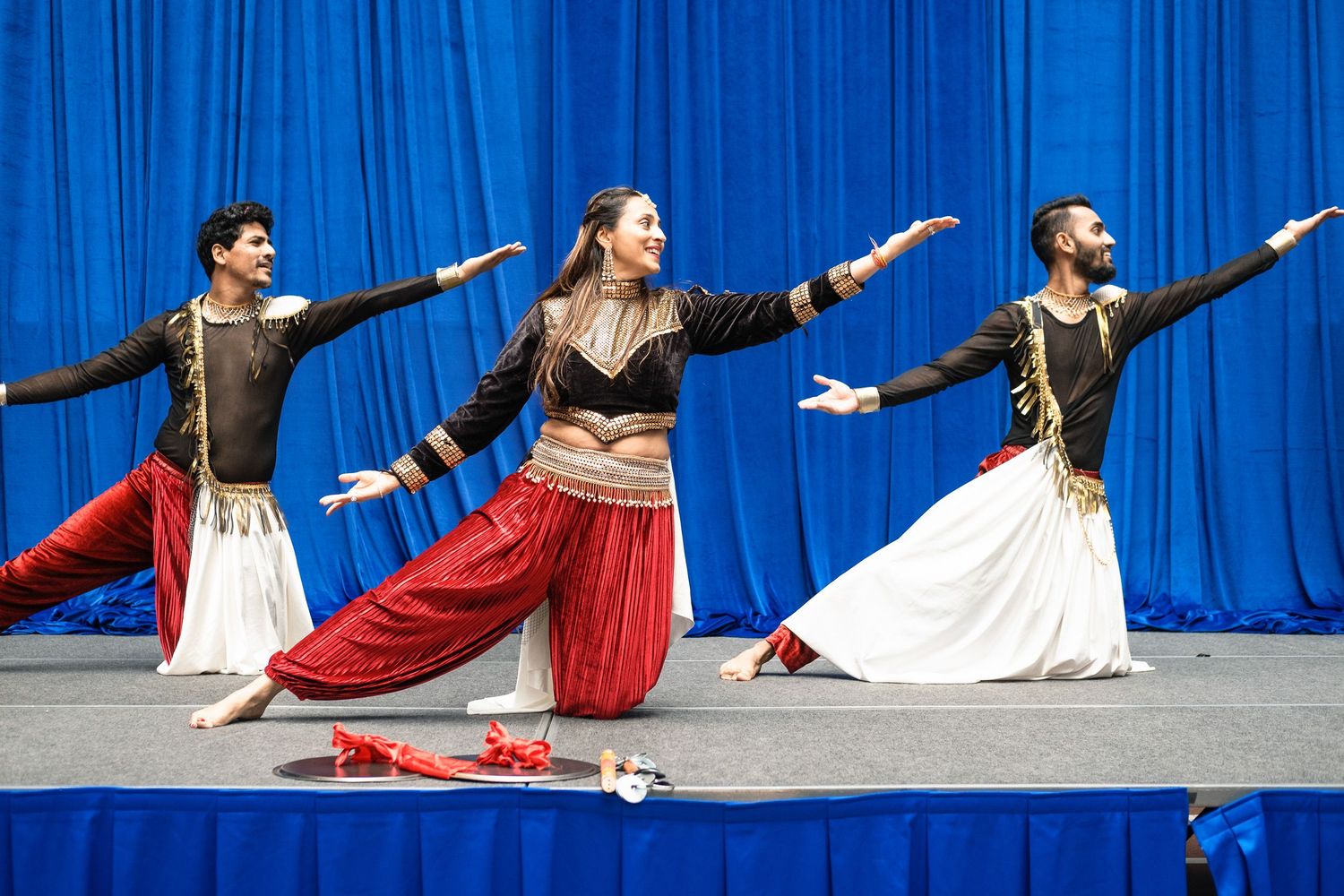 Three dancers perform classical Indian Kathak dance on stage with blue curtain backdrop, wearing traditional colorful costumes.