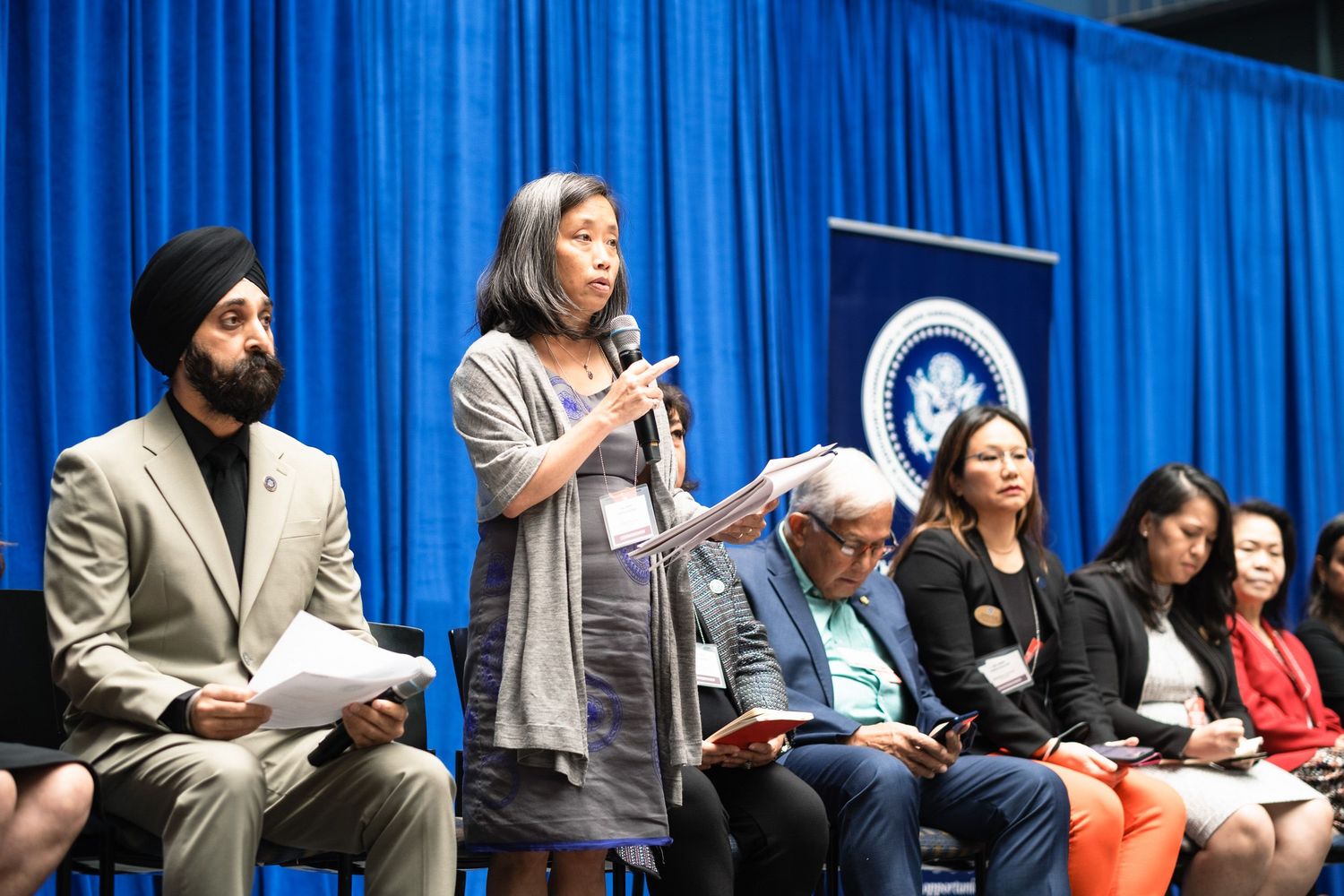 Diverse panel of speakers at a formal government or political event, with a woman addressing the audience at a podium.