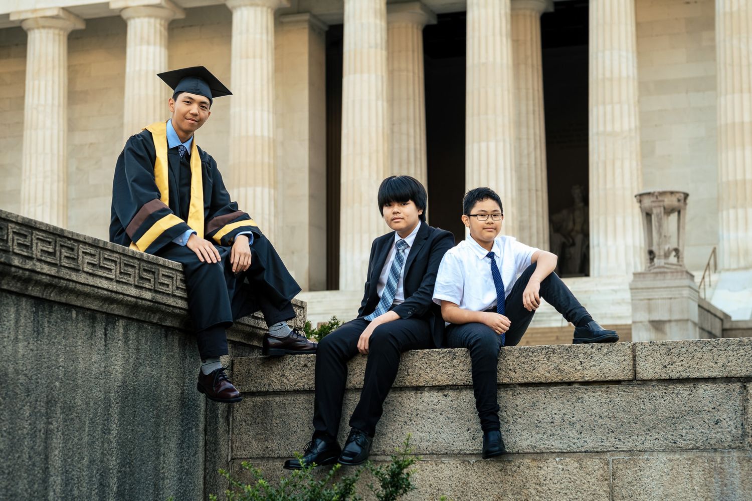 Three young Asian men in graduation and formal attire posing on steps of a classical building.