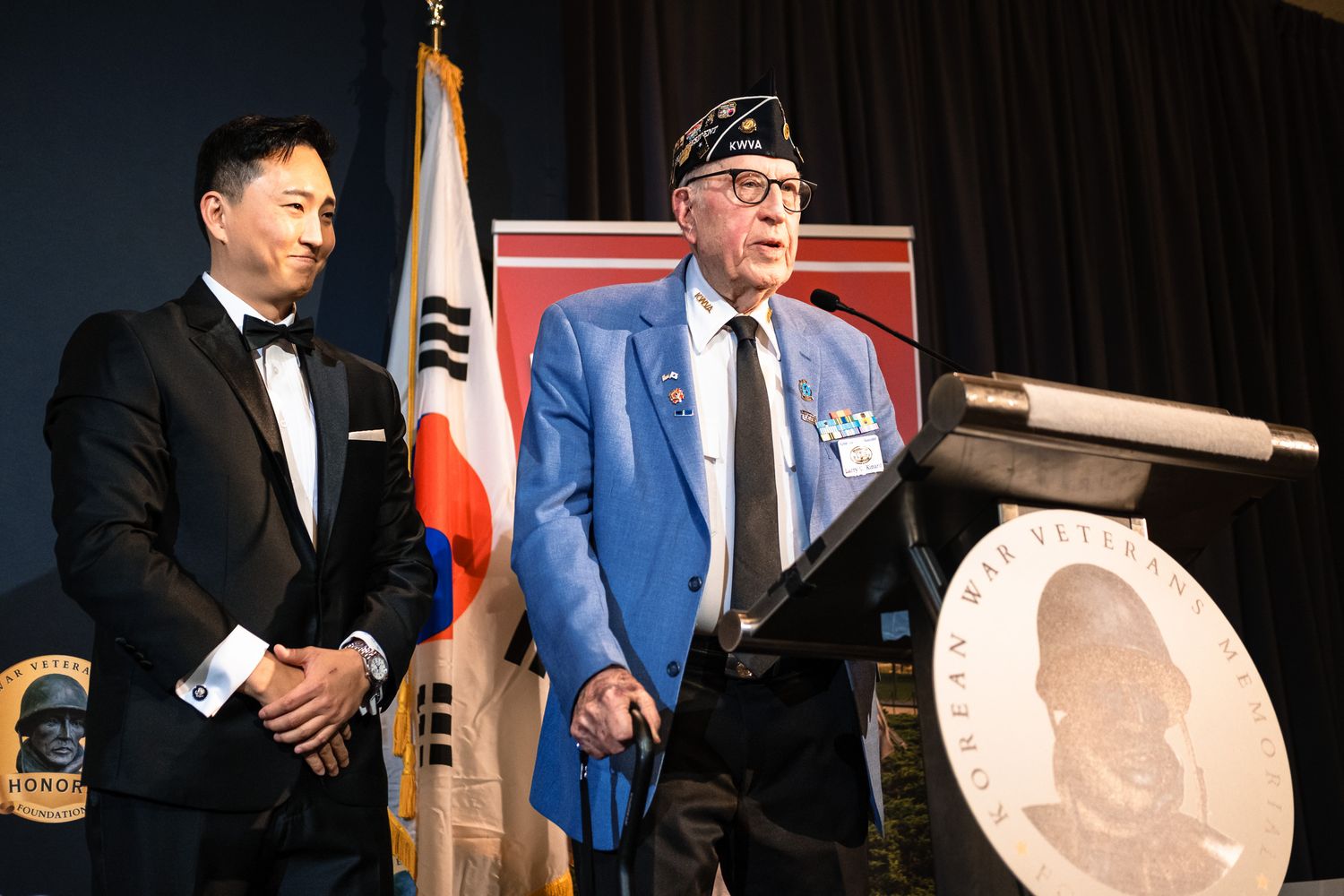 Korean War veteran in blue uniform speaks at podium beside man in tuxedo at formal ceremony.