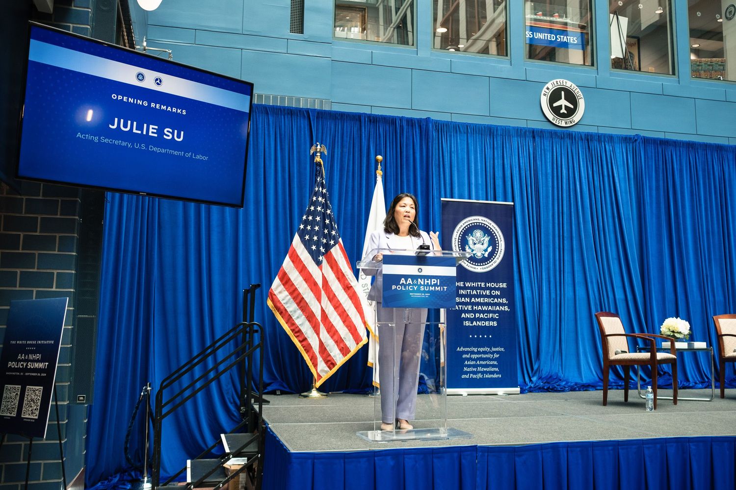 Julie Su speaks at a podium on a decorated stage with an American flag and official government seal backdrop.