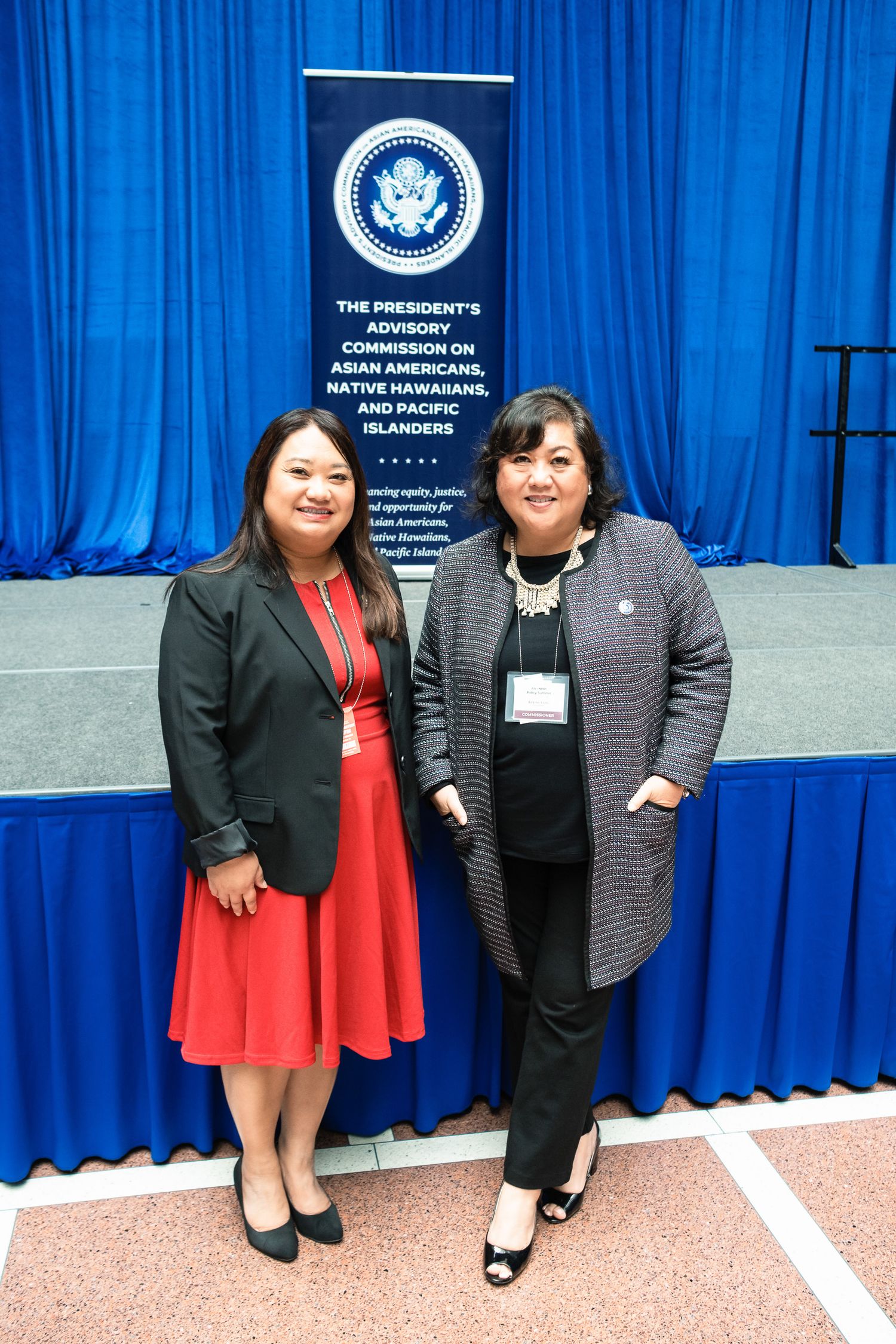 Two women pose together in front of the President's Advisory Commission on Asian Americans, Native Hawaiians, and Pacific Islanders banner.