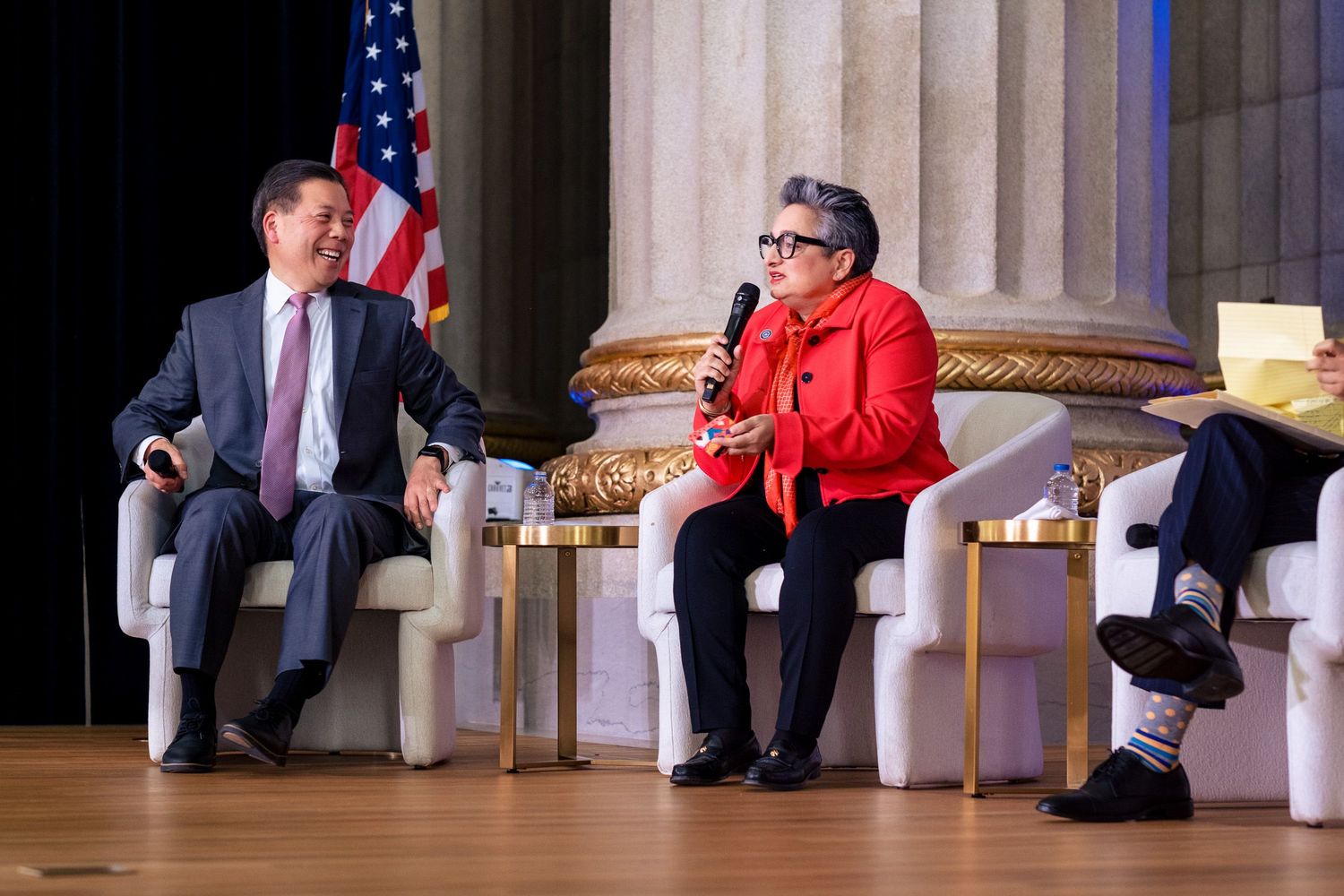 A man in a suit and a woman in red share a panel discussion at a formal event with an American flag backdrop.
