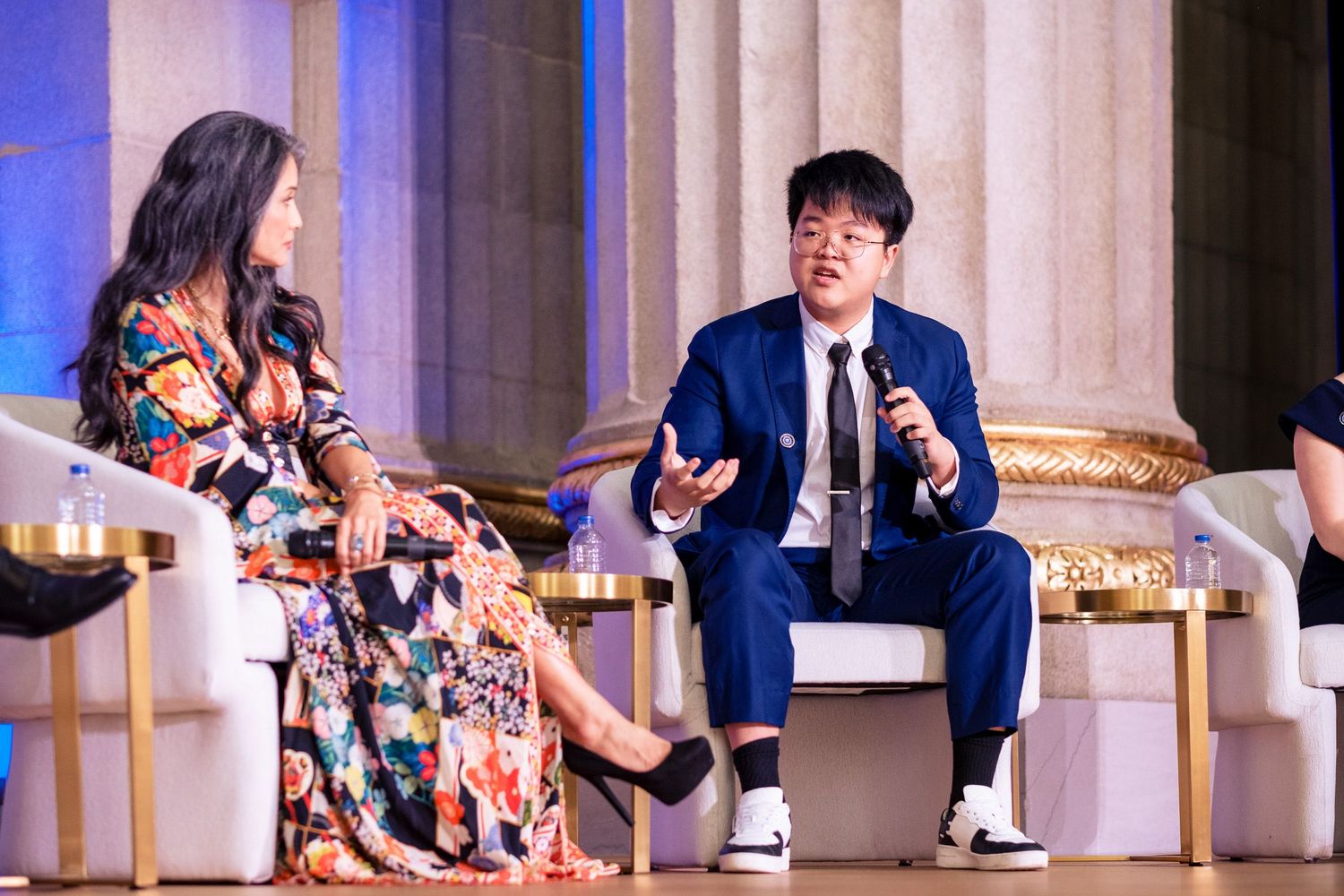 Young man in blue suit speaking on stage panel, gesturing expressively during discussion at formal event.