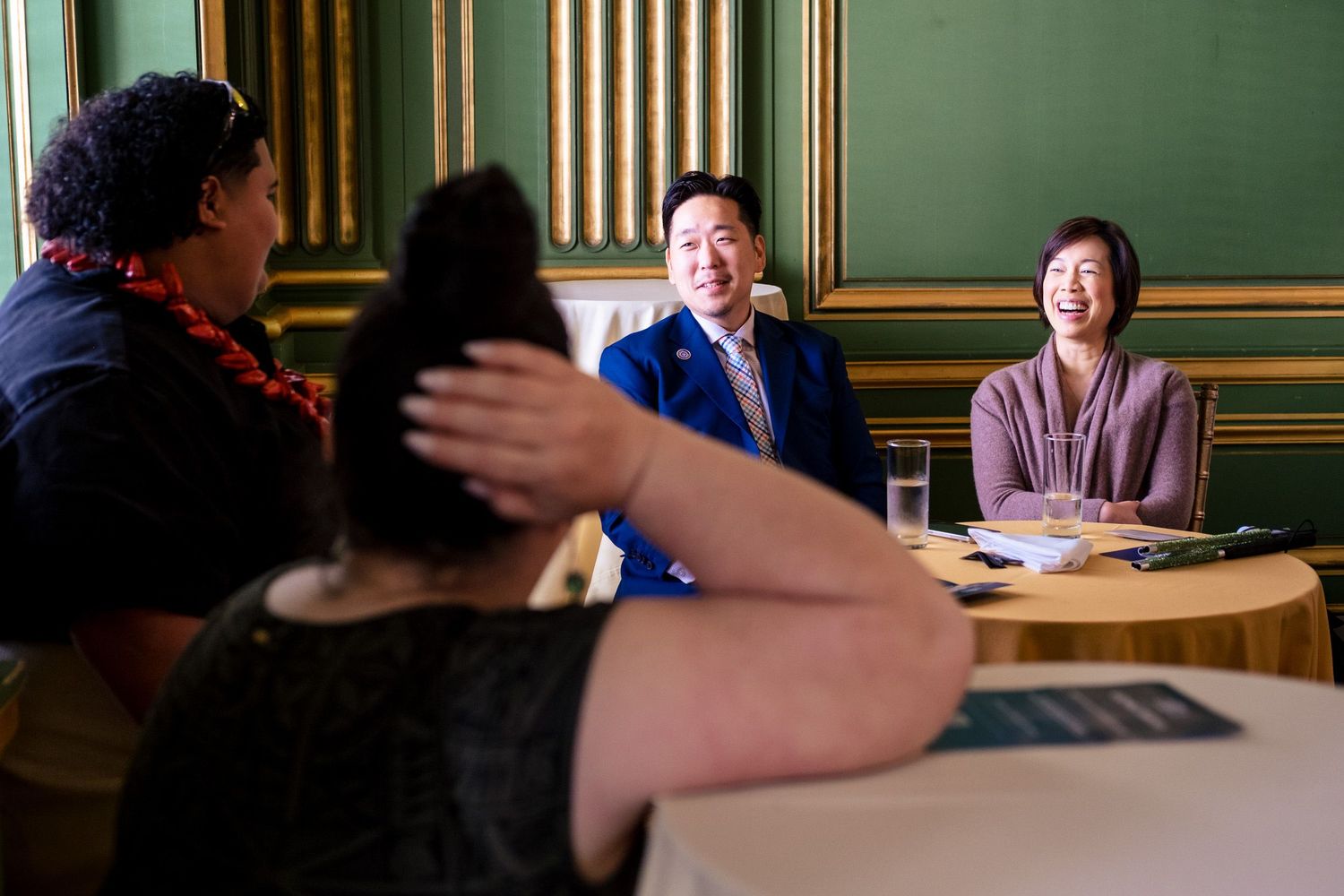 Two smiling professionals in a formal green-walled meeting room engaged in conversation with colleagues across a round table.