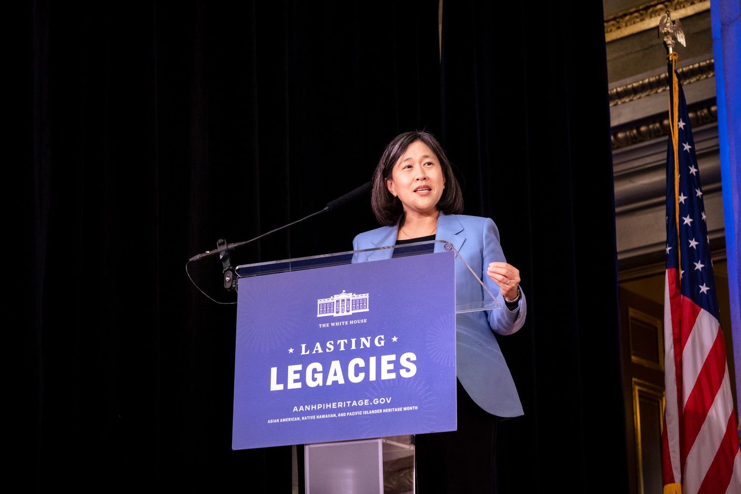 Woman in blue blazer speaks at podium with Lasting Legacies sign and White House logo at formal event.
