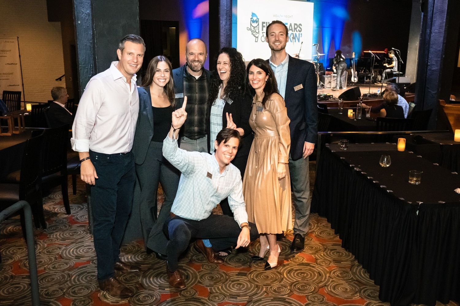 Group of seven professionals posing for a selfie at a corporate event in a hotel ballroom.