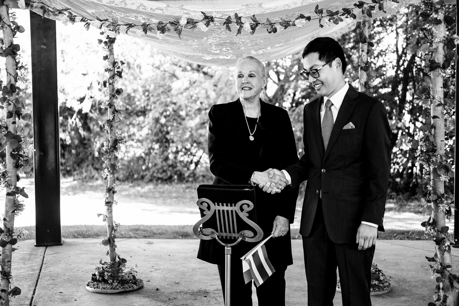 A black and white photo of a smiling officiant and groom standing together under a floral wedding arch.