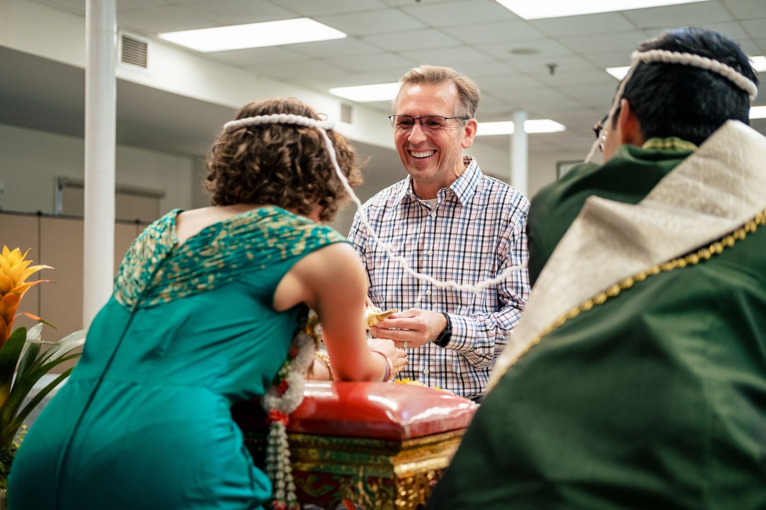 Smiling man presenting gift to couple during traditional Thai Buddhist wedding blessing ceremony indoors.