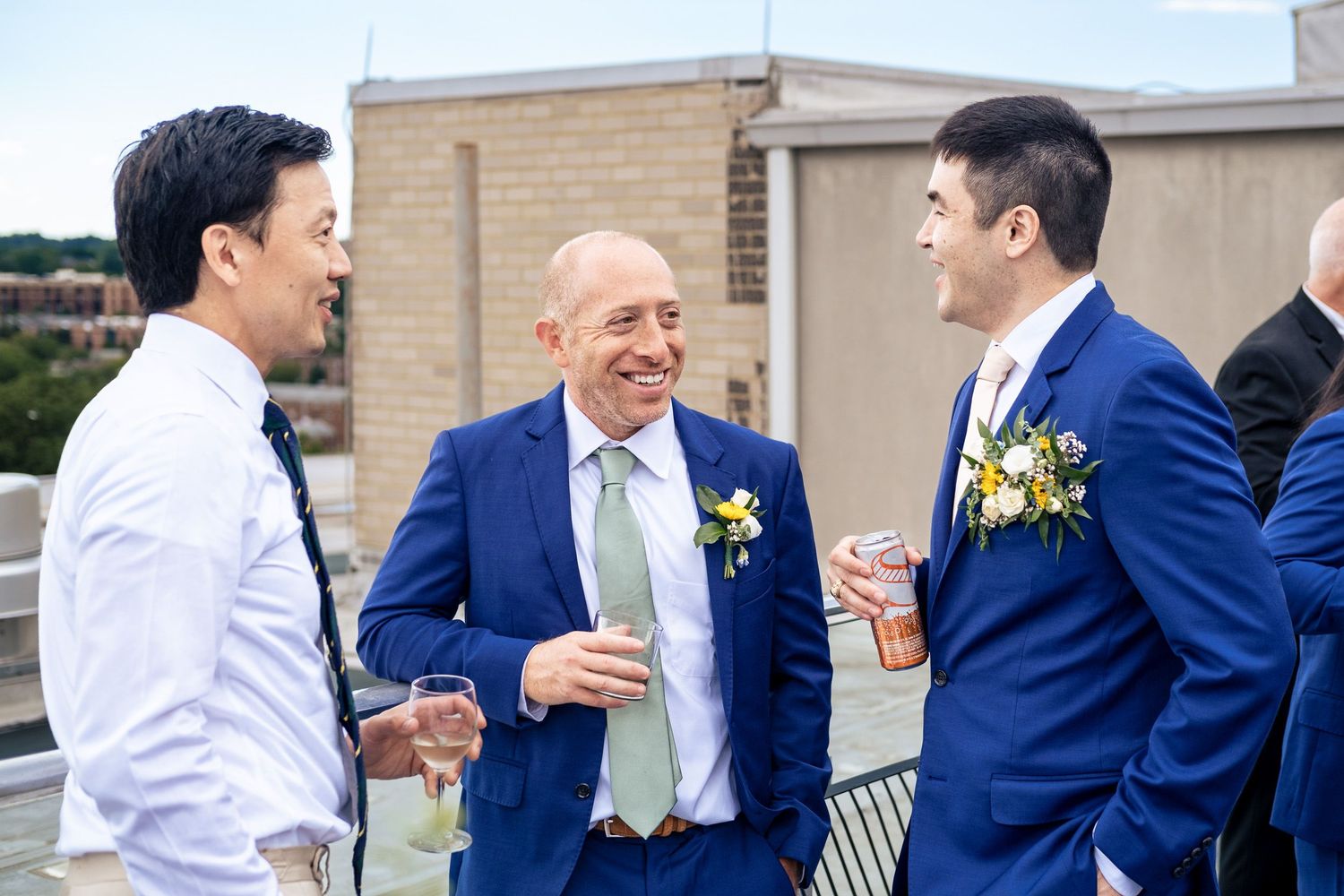 Three men in blue suits laugh and share drinks on a rooftop terrace at a wedding reception.