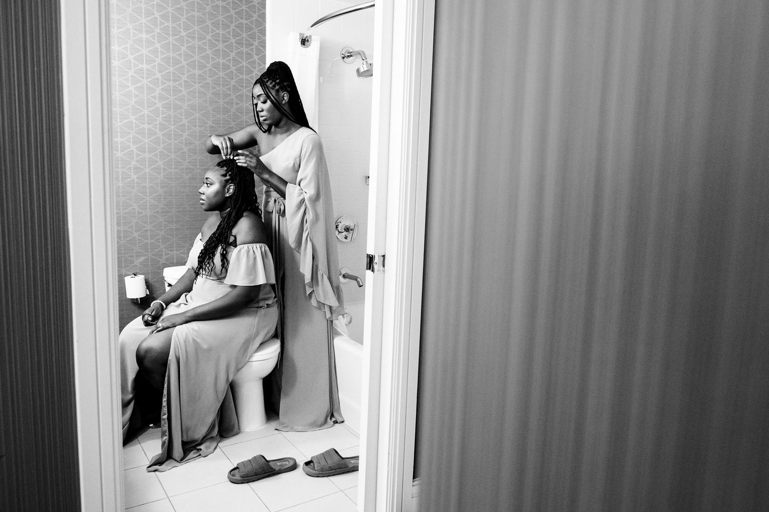 Black and white photo of woman styling seated bridesmaid hair in hotel bathroom mirror.