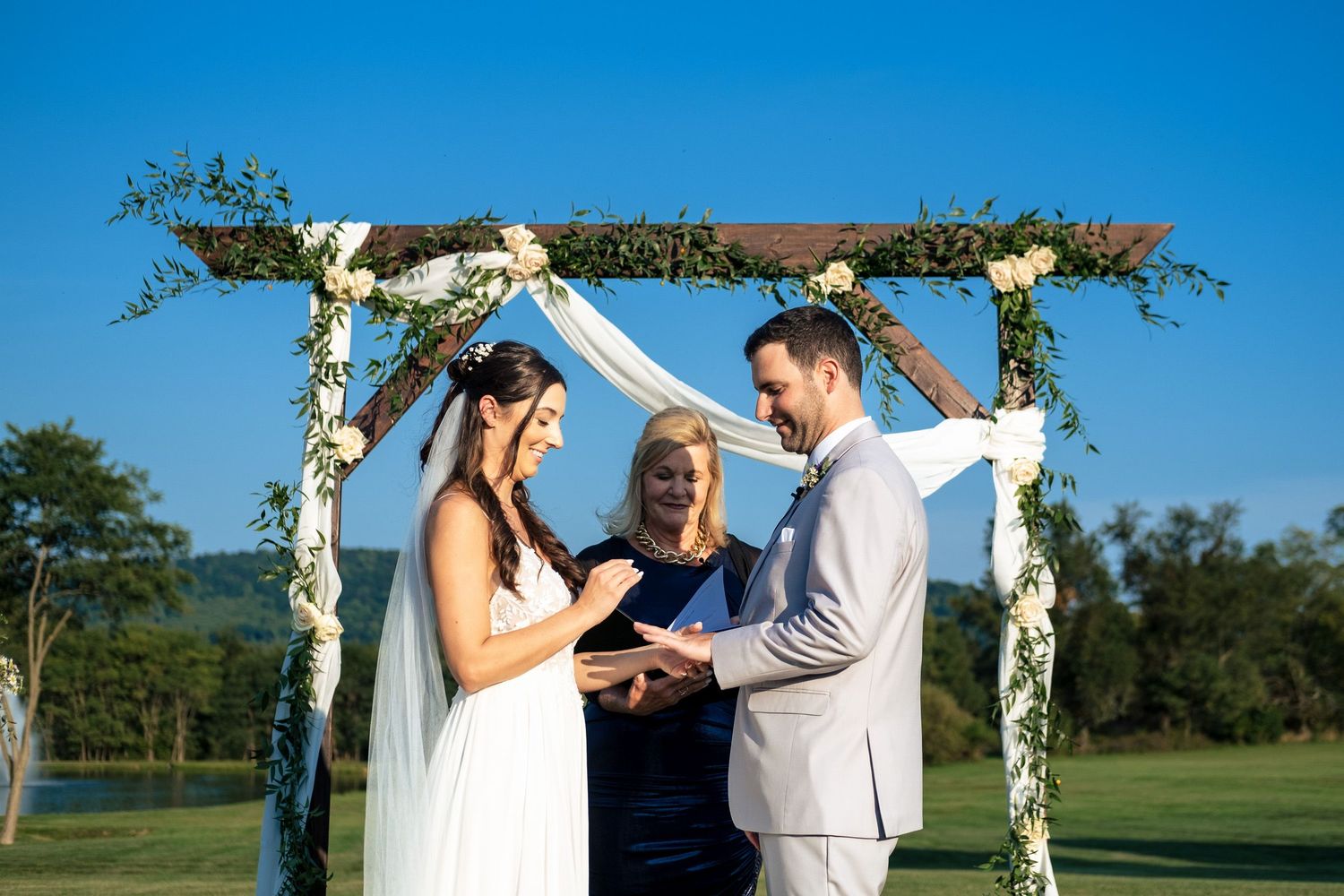 Bride and groom exchange rings under a rustic floral arch during a sunny outdoor wedding ceremony.