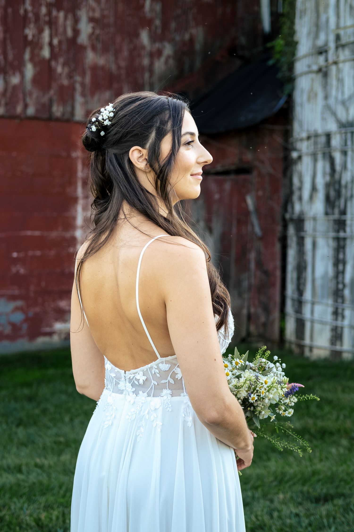 Bride in backless white lace wedding gown holds wildflower bouquet beside a rustic red barn.