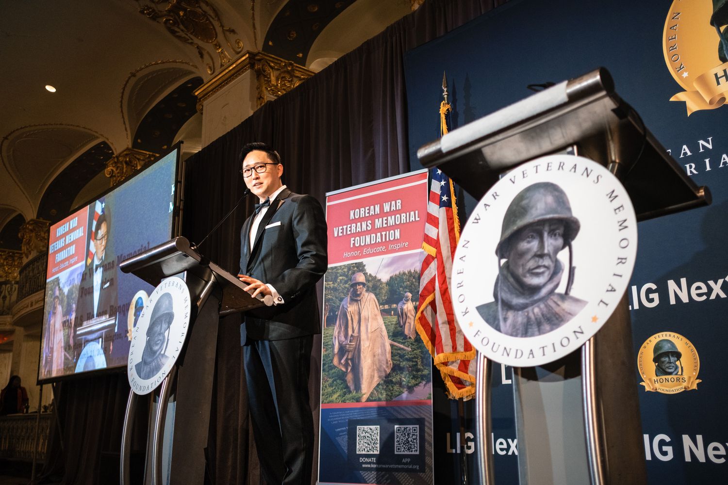 Speaker at podium during Veterans Memorial Foundation event with banners and display screens in background.