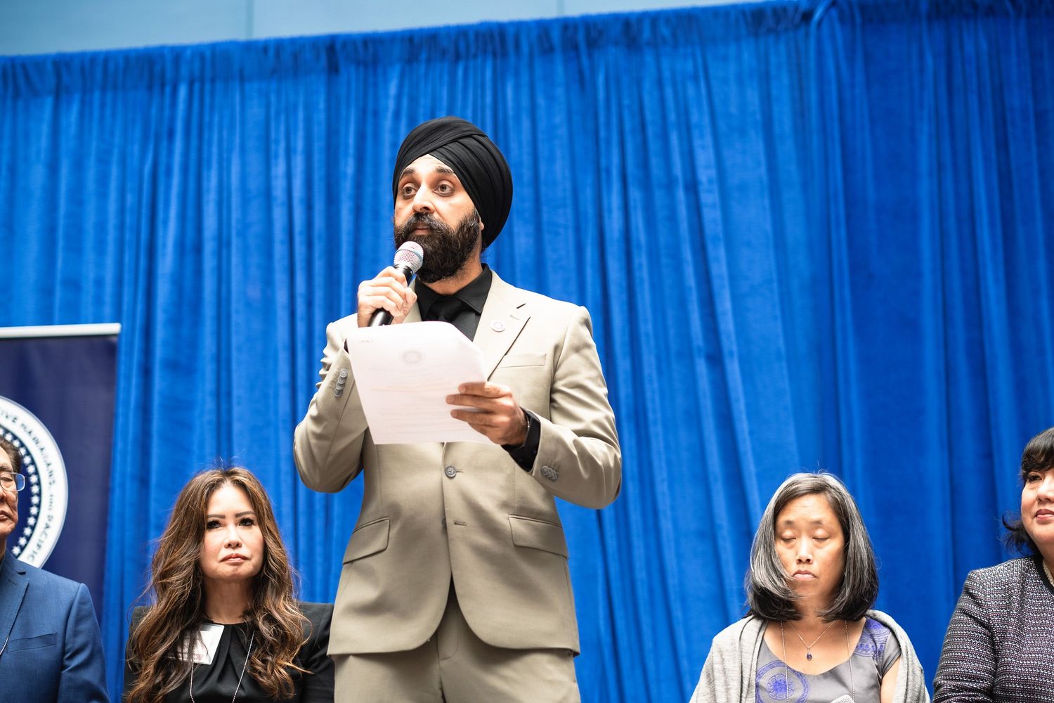Man in turban speaks at podium with microphone at official event with blue backdrop and seated audience.